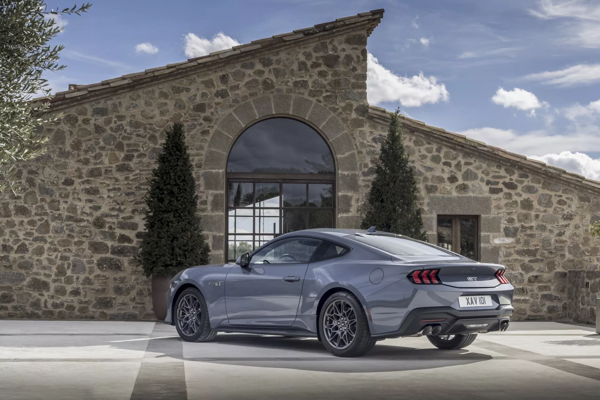 Ford Mustang coupé 2024 ukazující pohled zezadu a ze strany s LED zadními světly, dvojitým výfukem a velkými slitinovými koly na dlažebním nádvoří před rustikální budovou v jasný den, zdůrazňující jeho luxusní a výkonnostní vlastnosti.