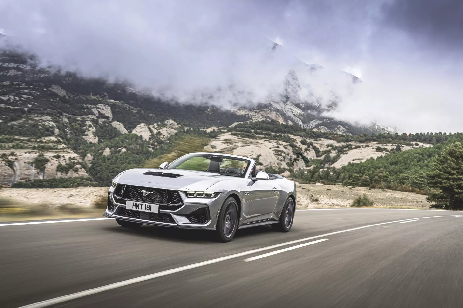 Ford Mustang Convertible 2024 v pohybu, ukazující ostré a úhlové designové prvky, včetně výrazné masky s charakteristickým vzorem, LED světlometů a tvarované kapoty, z nízkého úhlu zepředu ve třech čtvrtinách s horským pozadím