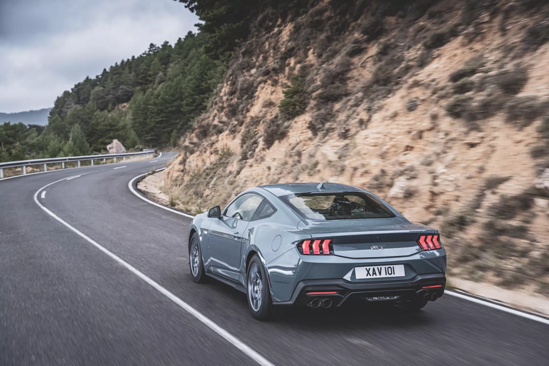 Stříbrné kupé Ford Mustang 2024 na serpentině: Zadní pohled 3/4 s důrazem na LED zadní světla, dvojité výfuky a aerodynamické prvky na pozadí krajiny