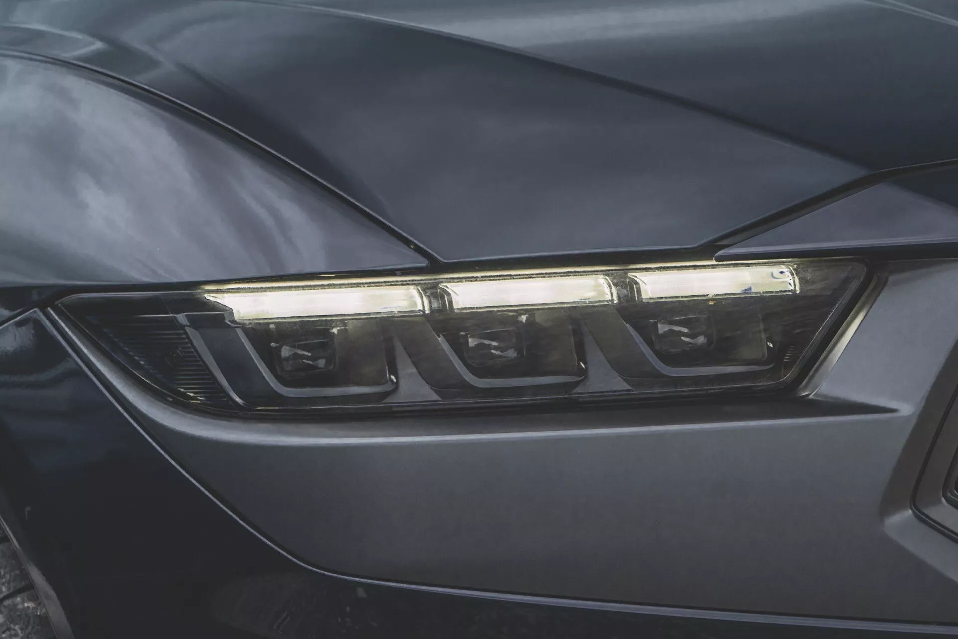 Detailní fotografie světlometu na straně řidiče Ford Mustang 2024 ukazující moderní technologii LED nebo HID a aerodynamický design v tmavém provedení.