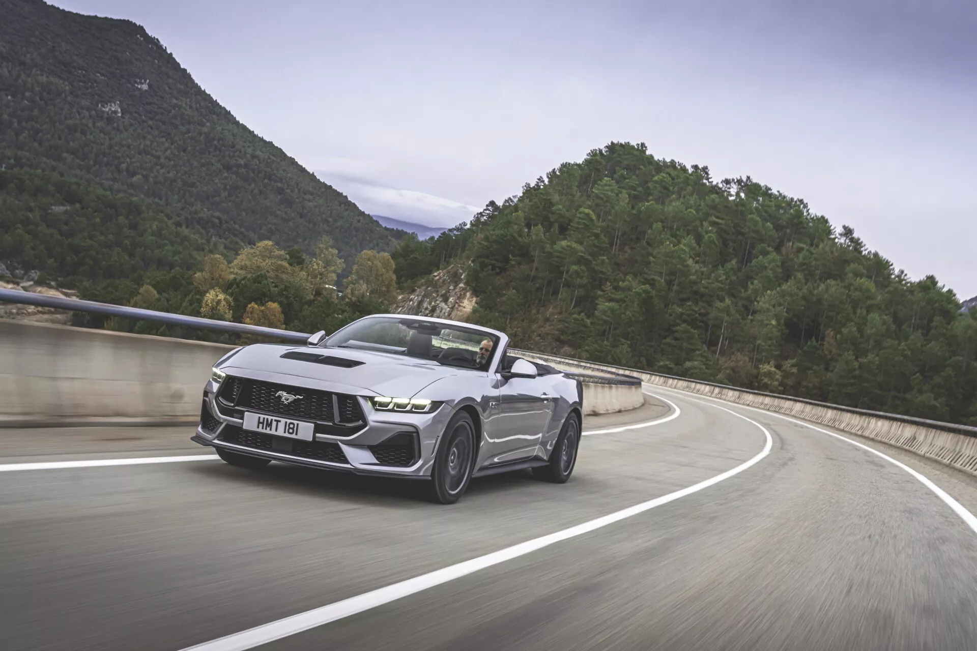 Ford Mustang 2024 v pohybu na horské silnici, prezentující jeho moderní design muscle caru s ikonickým emblémem koně ve skoku, agresivní přední částí s úzkými světlomety, sportovním nárazníkem, profilovanými liniemi kapoty, charakteristickými slitinovými koly a skládací střechou v metalickém provedení.