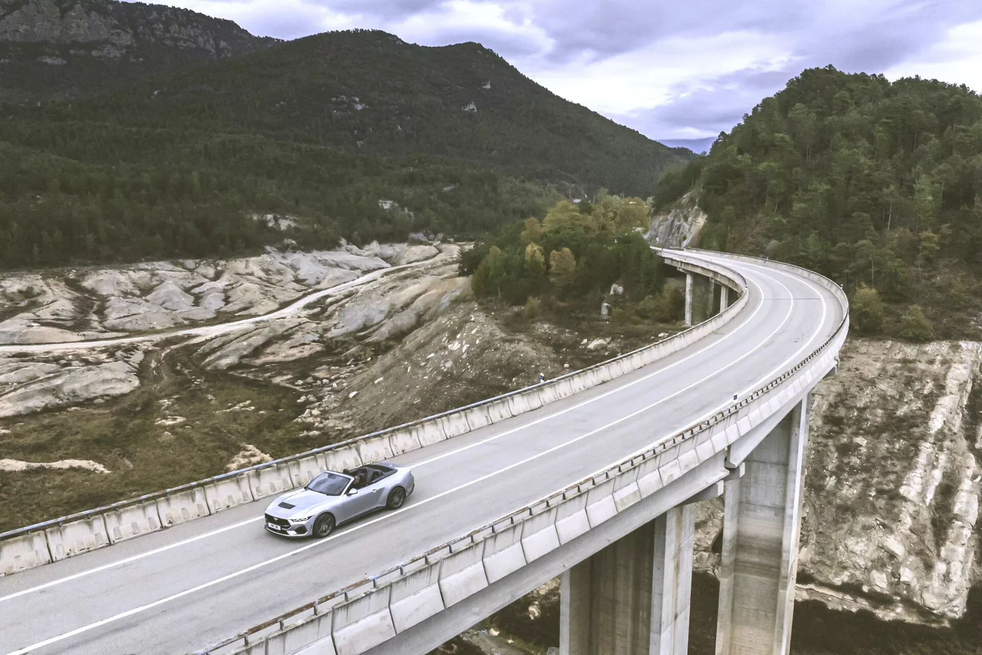 Ford Mustang 2024 cestující po mostě v kopcovité krajině, zachycen z vysokého úhlu ukazující přední levou stranu a pohled zeshora se zaměřením na kovový lak a designové prvky jako přední mřížka a slitinová kola pod přirozeným světlem