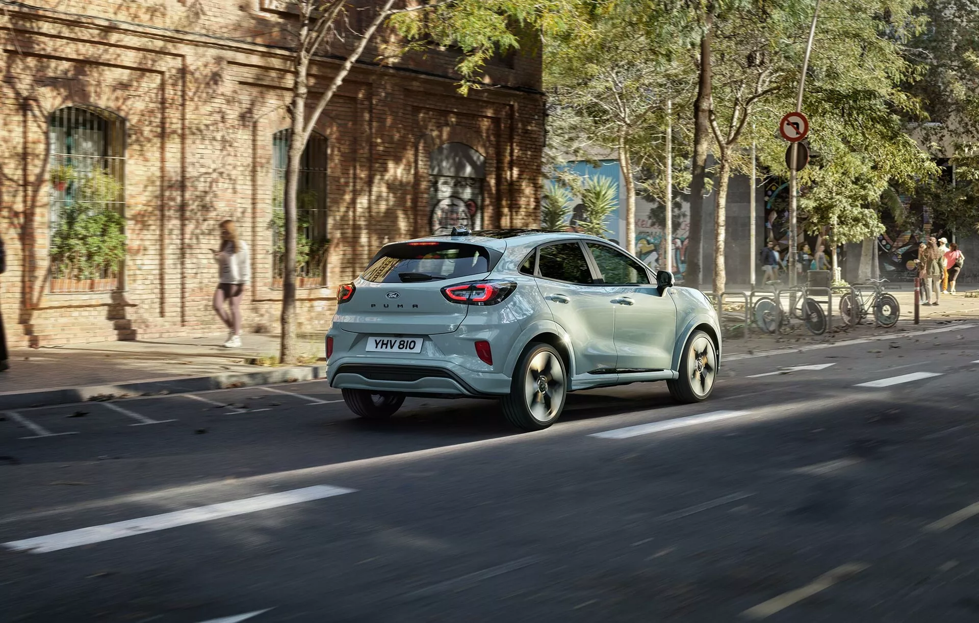 Ford Puma 2024 (II-facelift) světle zbarvený hatchback v pohybu na městské ulici z trojčtvrtě zadního úhlu ukazující zadní světla, zadní okno, boční dveře a slitinová kola