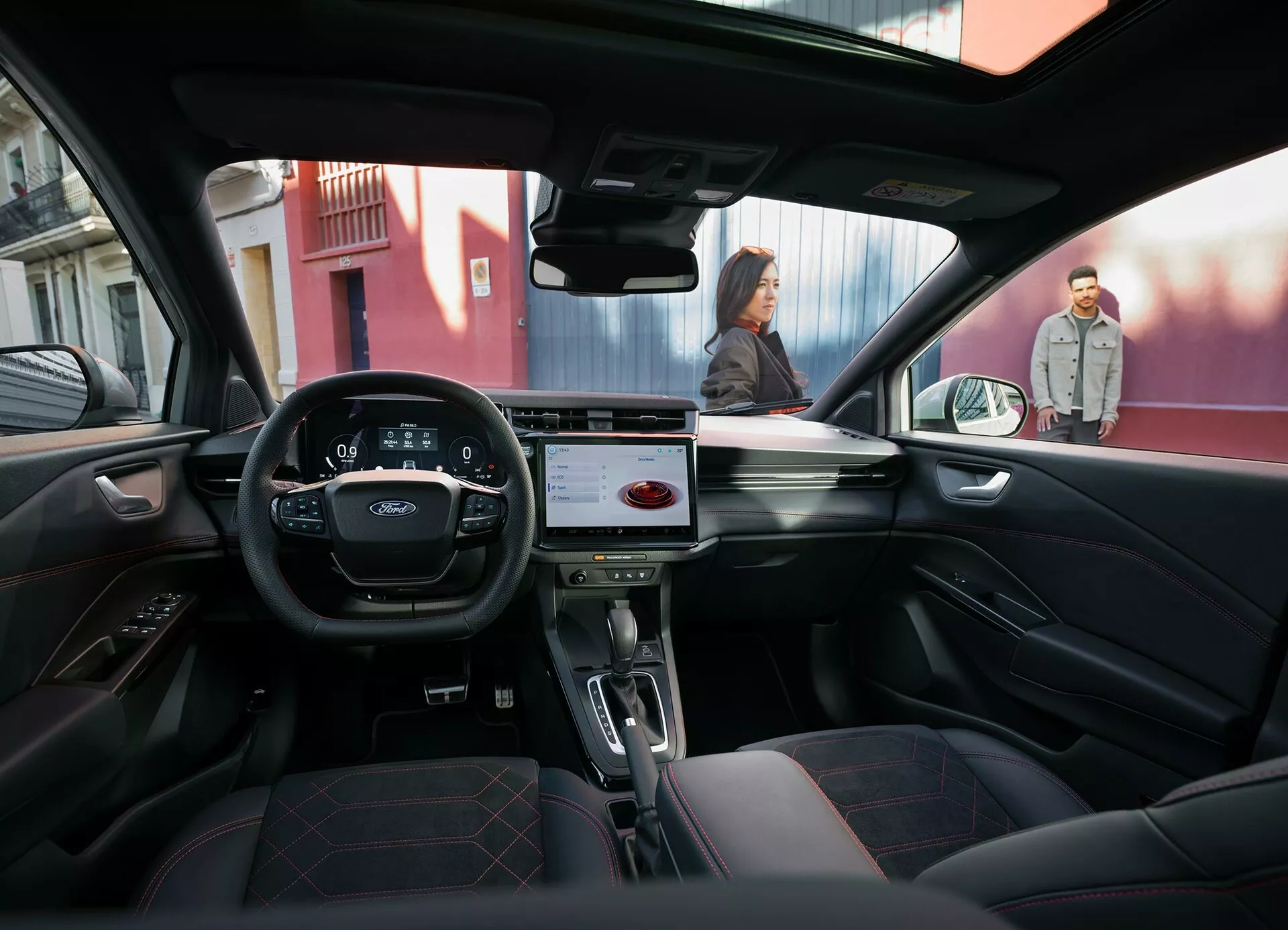 Pohled z interiéru z zadního sedadla vozu Ford Puma 2024 ukazuje palubní desku s dotykovým informačním systémem, přední sedadla z kůže, volant s ovládacími prvky a městský výhled zvenčí přes čelní sklo