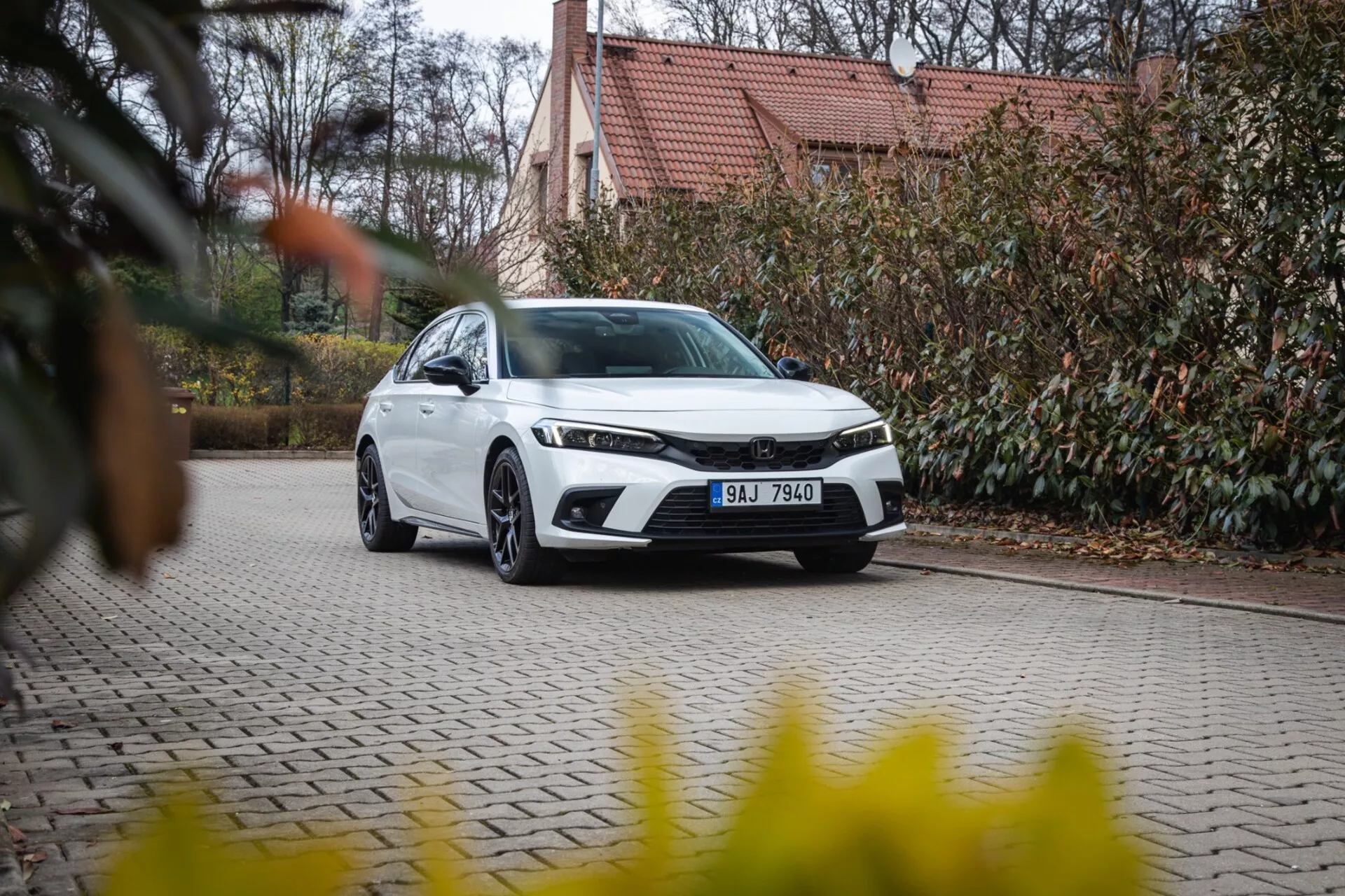 Moderní sedan Honda Civic 2022 zaparkovaný na dlážděné příjezdové cestě se zahradní úpravou a budovou v pozadí, představující přední mřížku, LED světlomety a elegantní aerodynamický design