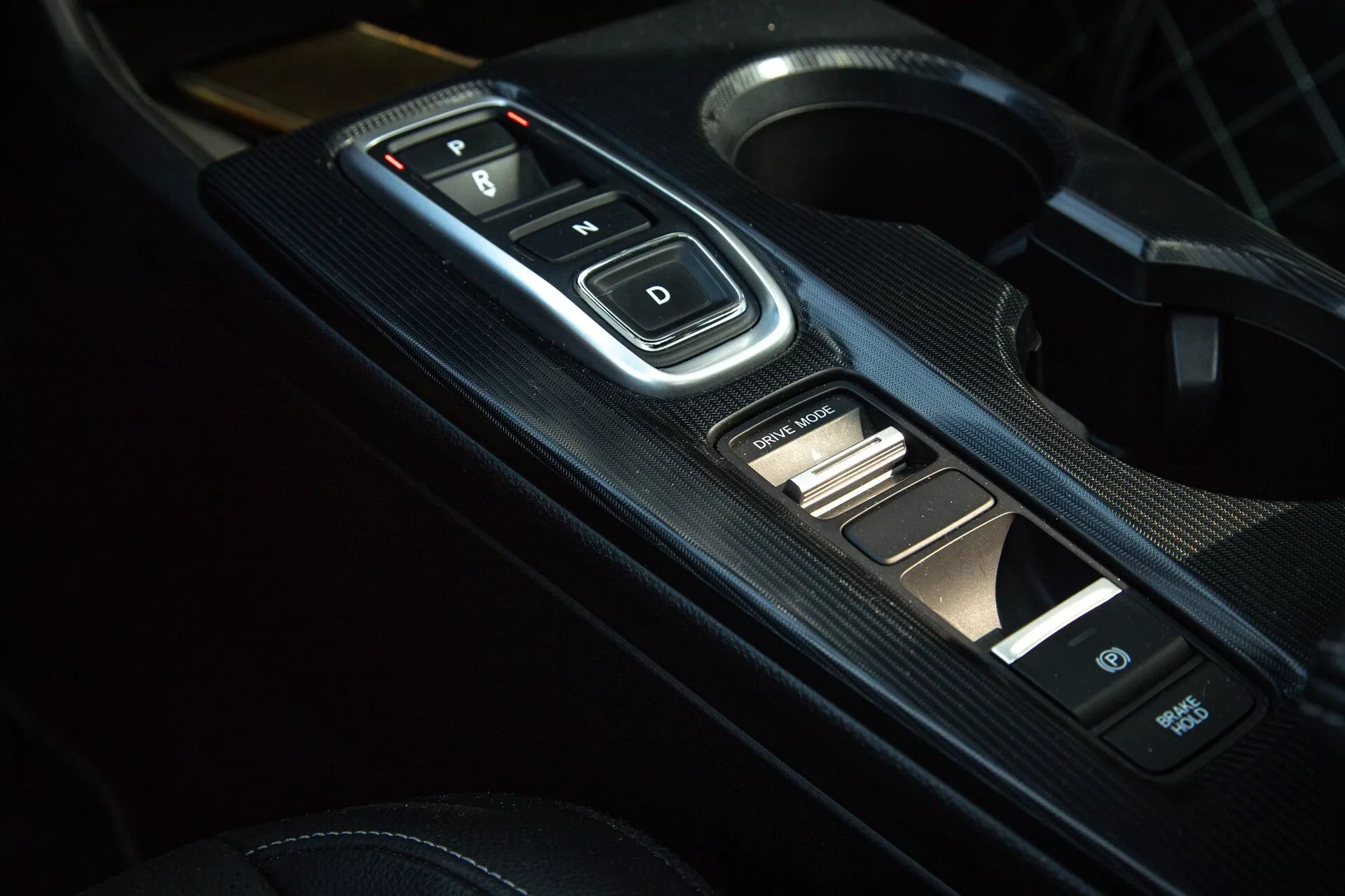 Detail interiéru Hondy Civic 2022 se zaměřením na středovou konzolu s elektronickým voličem rychlostí s tlačítky 'P, R, N, D', panelem s texturou z uhlíkových vláken a doplňkovými ovladači, předvádějící moderní automatickou převodovku a sportovní estetiku