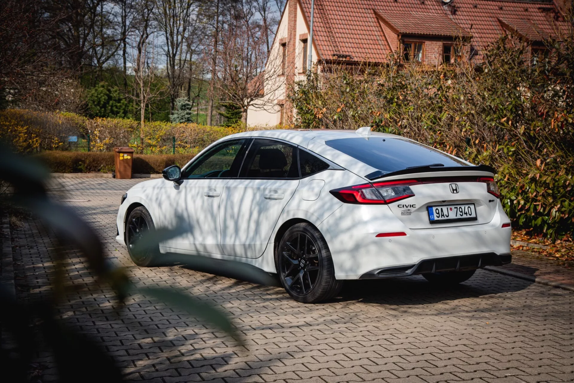 bílý Honda Civic z roku 2022 s černými vícepaprskovými koly a zadním spoilerem, zaparkovaný na dlažbě s podzimním listím a obytným domem na pozadí