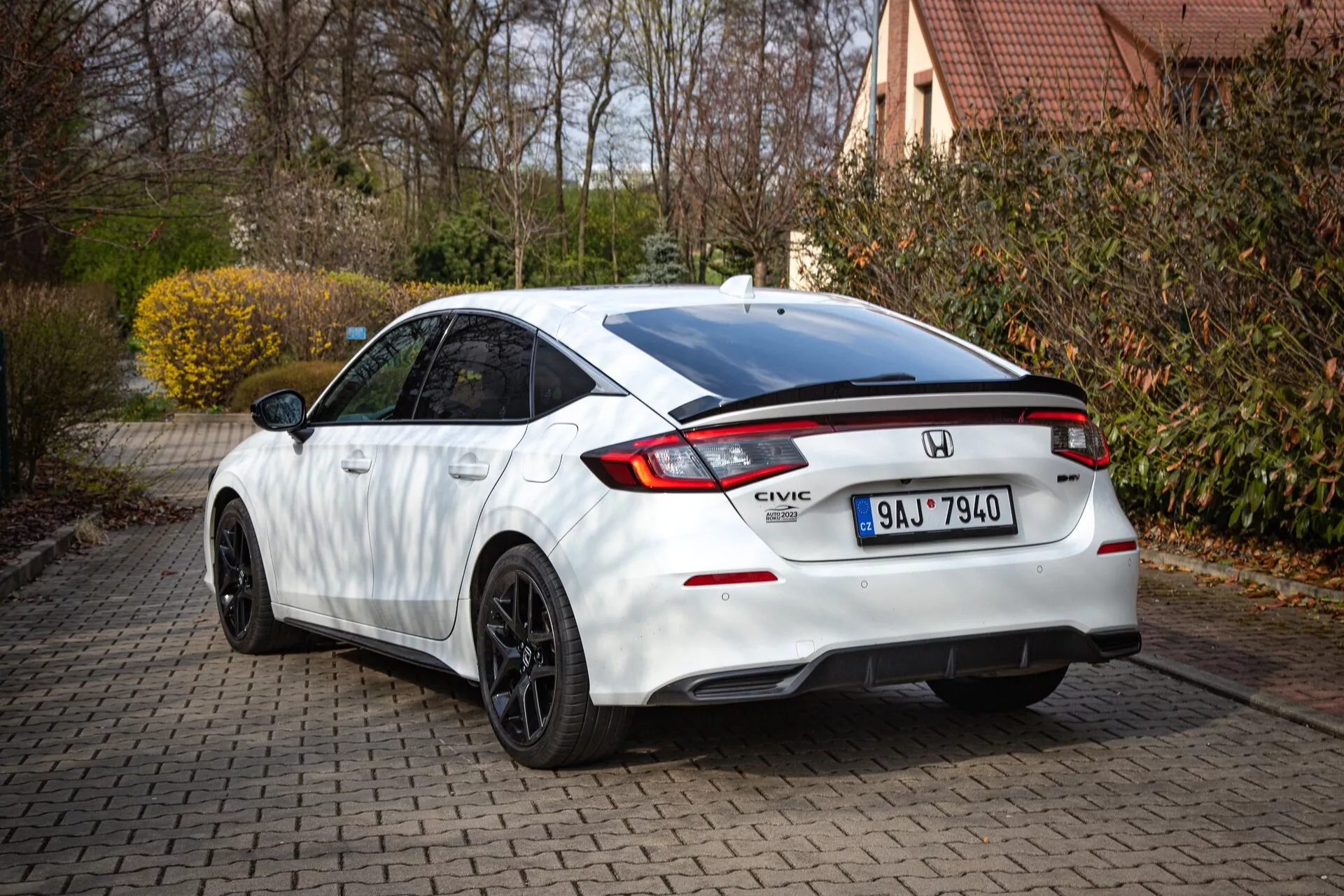 Bílý Honda Civic 2022 zobrazující zadní a částečný boční profil na cihlové příjezdové cestě, se svažující se střechou, integrovaným spoilerem, LED brzdovým světlem, charakteristickými zadními světly a sportovním nárazníkem s difuzorem