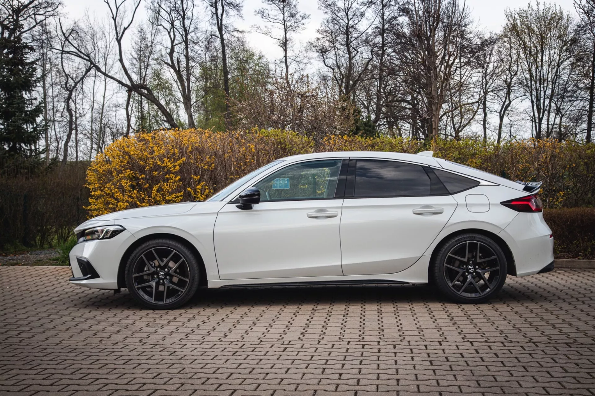 Bílý čtyřdveřový sedan Honda Civic 2022 zaparkovaný na vydlážděném prostoru s propojenými cihlami, zobrazující boční profil se sportovním designem a moderními LED světlomety, zachycený z mírně zvýšeného úhlu pro zvýraznění celého bočního pohledu včetně tónovaných oken, sportovních slitinových kol a technologických prvků, jako jsou integrované směrové signály v bočních zrcátkách.