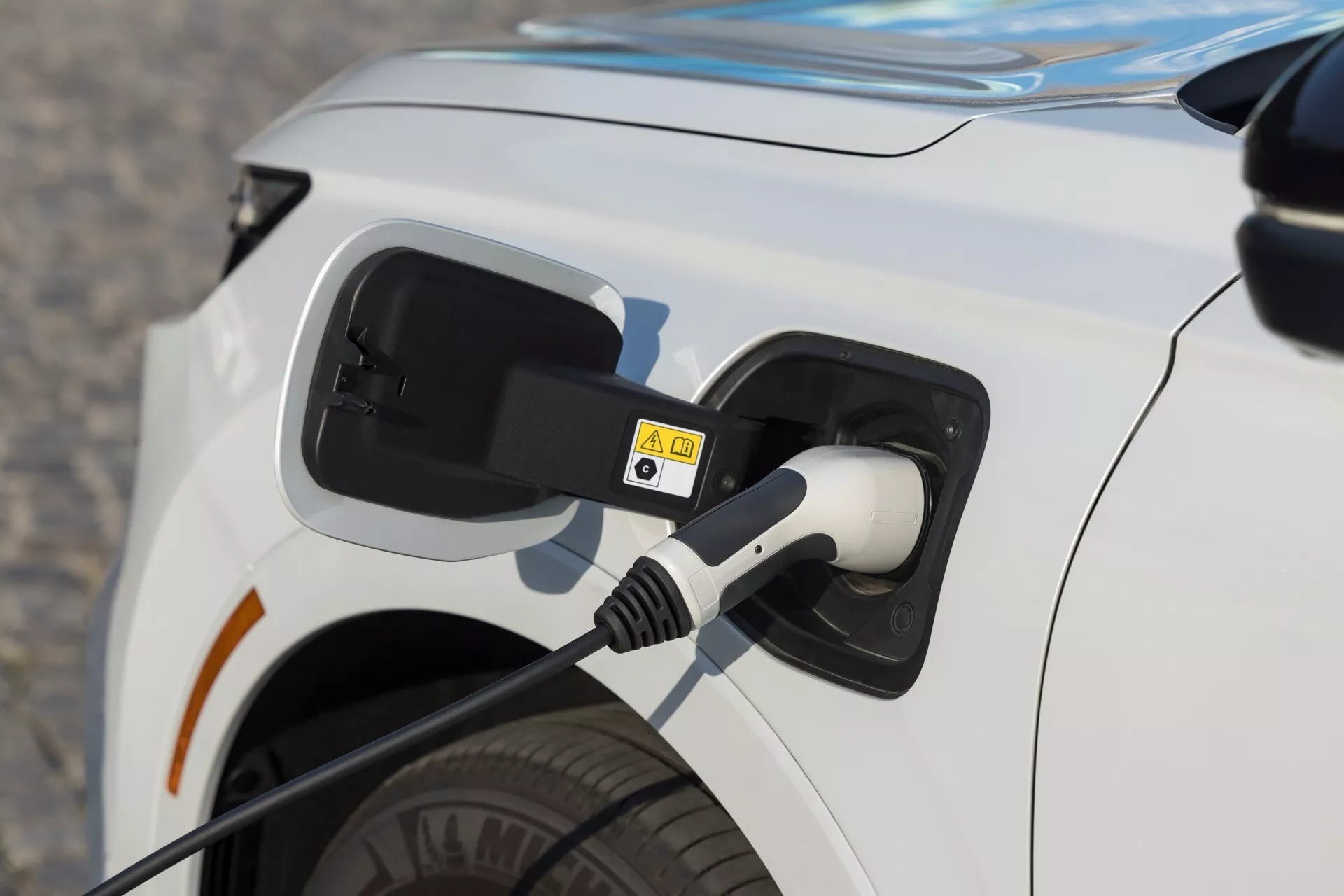Detailní záběr na nabíjecí port na levé zadní části bílého elektromobilu Honda CR-V 6. generace z roku 2023 s připojeným nabíjecím kabelem a varovnými štítky vysokého napětí, zdůrazňující elektrickou a ekologickou technologii vozidla.
