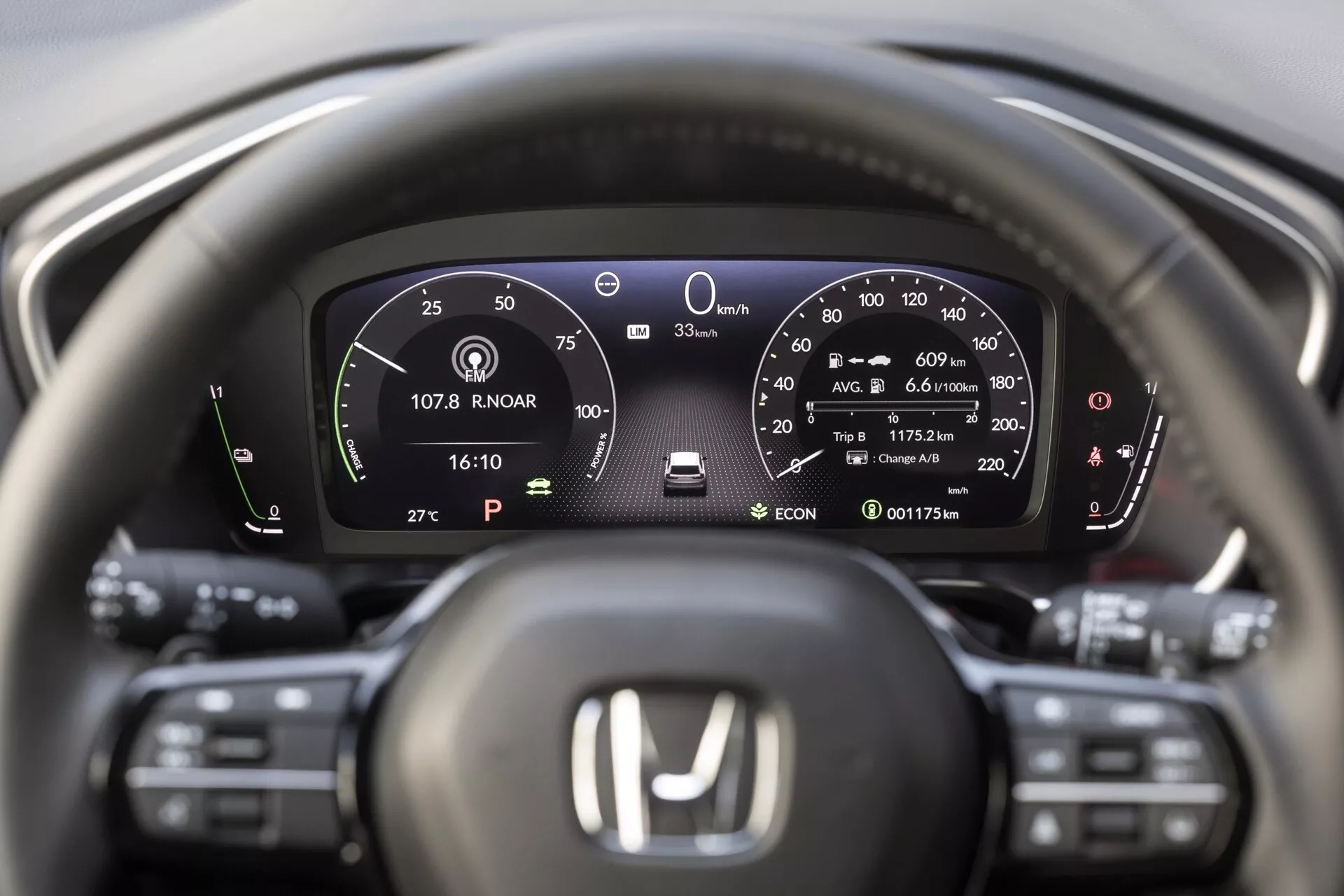 Interiérový pohled na Honda CR-V 6. generace z roku 2023 zaměřený na digitální přístrojový panel a oblast volantu, zobrazující rychlost vozidla, indikátor ekologické jízdy, ukazatel paliva, režim parkování, systém parkovací brzdy, indikátor dojezdu, čas, venkovní teplotu, počítač cesty a ovládání asistence řidiče.