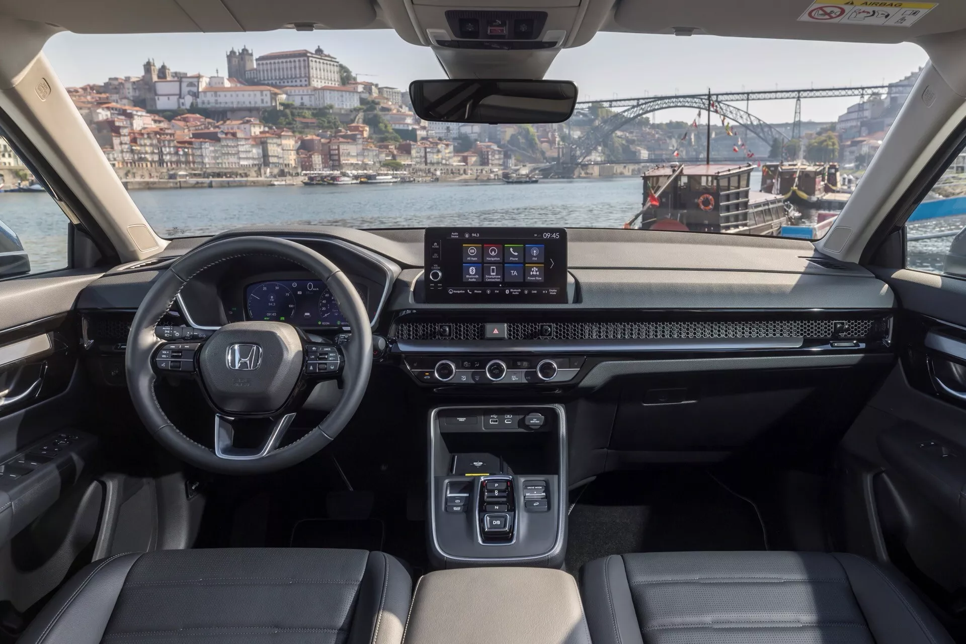 Interiér Hondy CR-V 2023 z pohledu ze zadního sedadla s širokoúhlým infotainmentem, ovládáním klimatizace a koženými sedadly s osvětlením denním světlem a městskou krajinou přes čelní sklo
