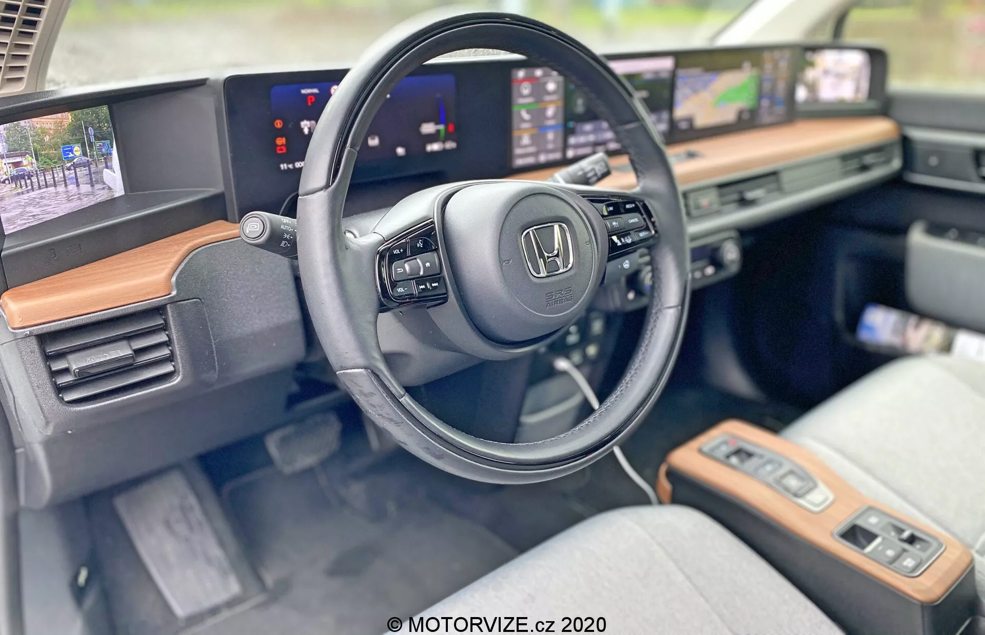 Interiér modelu Honda E 2020 zaměřený na stranu řidiče s důrazem na volant a moderní palubní desku. Makro záběr zvýrazňuje logo Honda na volantu, který má namontované ovládání pro multimédia a tempomat. Prodloužené, souvislé obrazovky slouží jako digitální přístrojová deska zobrazující rychlost a infotainment systém s navigační mapou. Viditelný dřevěný dekor přidává dotek luxusu a světlejší látkové potahy sedadel naznačují pohodlí a styl. Pokročilé funkce jako dotykové panely, tlačítko na start a bezklíčové zapalování podtrhují technologicky vybavenou kabinu.