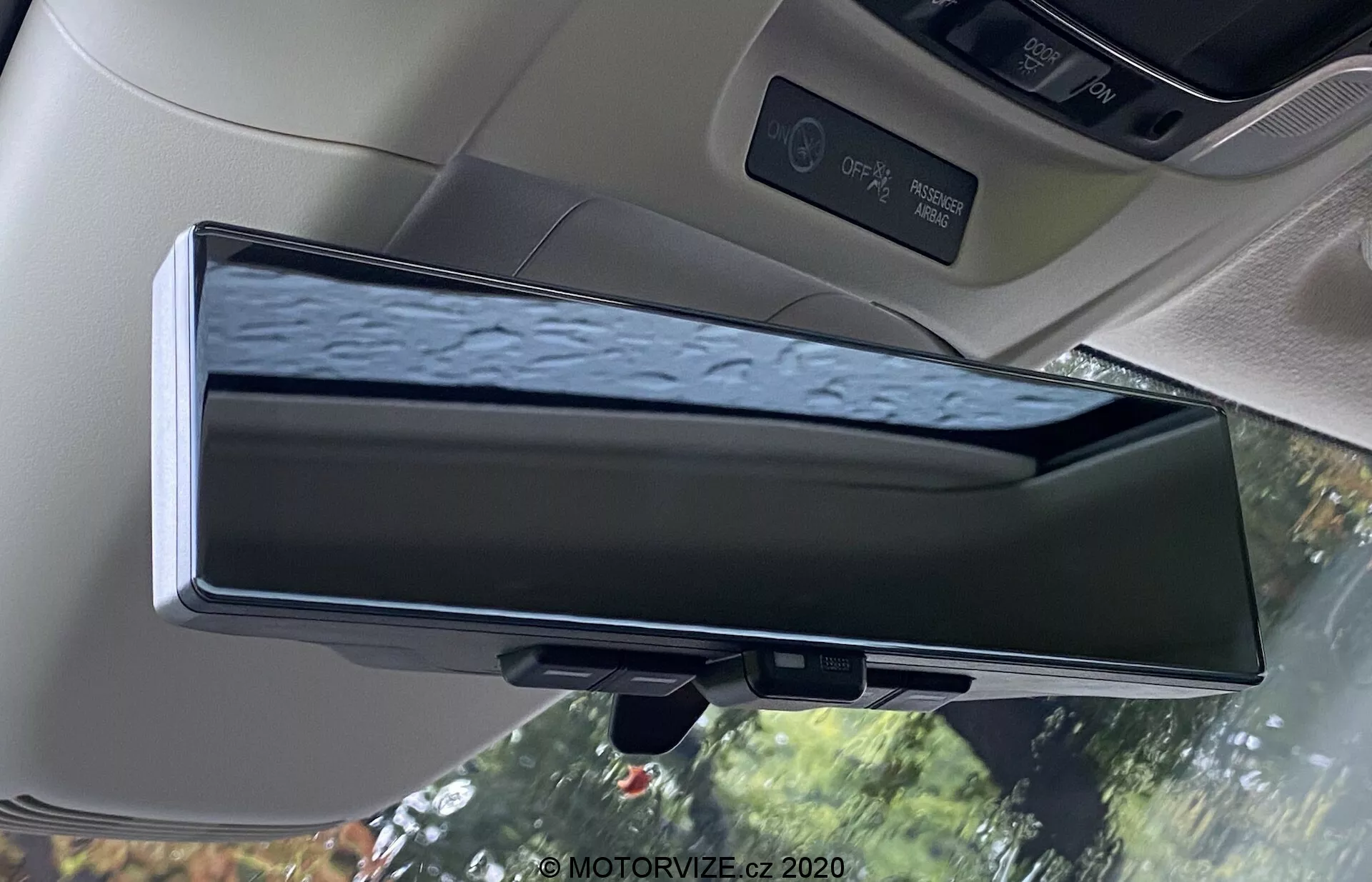Pohled na interiér vozu Honda E 2020 s digitálním zpětným zrcátkem. Elektronické zařízení je umístěno v tradičním krytu zpětného zrcátka a zobrazuje živý přenos z kamery na zadní straně vozu. Zrcátko zobrazuje odrazy stromů s kapkami vody a nabízí lepší viditelnost ve srovnání s tradičními zrcátky. Obrázek zachycuje stropní obložení vozu, kabinová světla, sluneční clonu a ovládací panel střešního okna, a zdůrazňuje technologii pokročilých asistenčních systémů řidiče (ADAS).