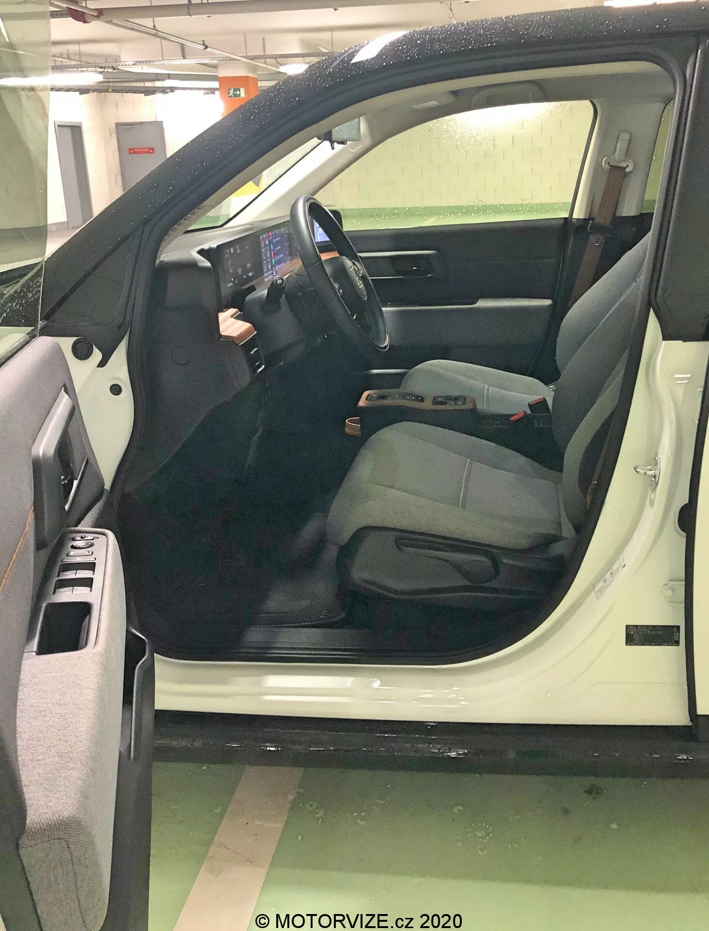 Detailní pohled na otevřené dveře na straně řidiče automobilu z mírně zvýšeného úhlu, zaměřený na moderní, technologicky zaměřený interiér. Velká prominentní centrální dotyková obrazovka, minimální fyzická tlačítka a ovládací prvky namontované na volantu zdůrazňují pokročilé informační a ovládací systémy. Sedadlo řidiče naznačuje komfort s nastavitelnou opěrkou hlavy. Detaily zahrnují úložné přihrádky ve dveřích, mřížku reproduktorů, ovladače elektrických oken, bezpečnostní pásy, nastavovací páčky a koberečky, což signalizuje novější, dobře udržovaný model.