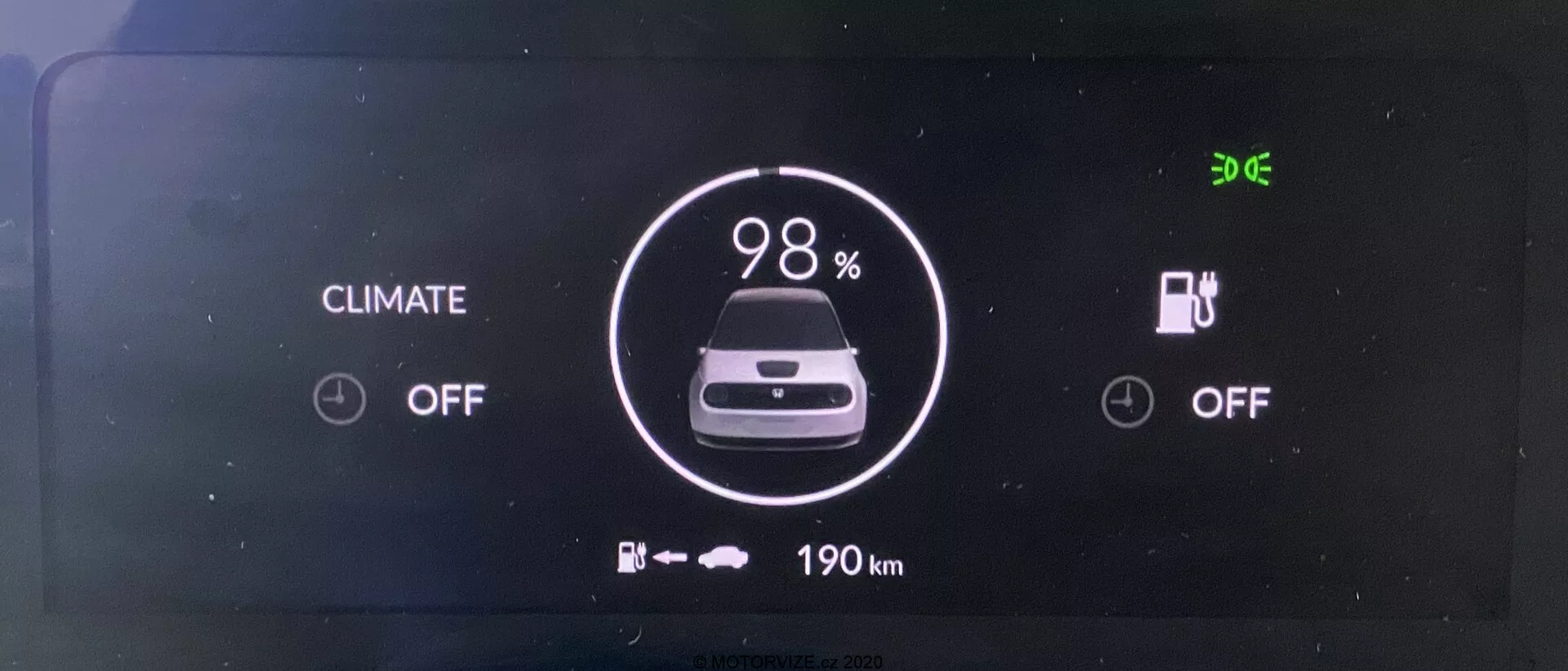 Digitální přístrojová deska elektrického vozidla Honda e 2020. Obrazovka zobrazená přímo čelně jasně ukazuje indikátor baterie na 98% nabití, ikonu auta a pod ní '190 km' zbývajícího dojezdu. Stav klimatizace a nabíjení je vypnutý. V levém horním rohu je zobrazena čas '9:06'.