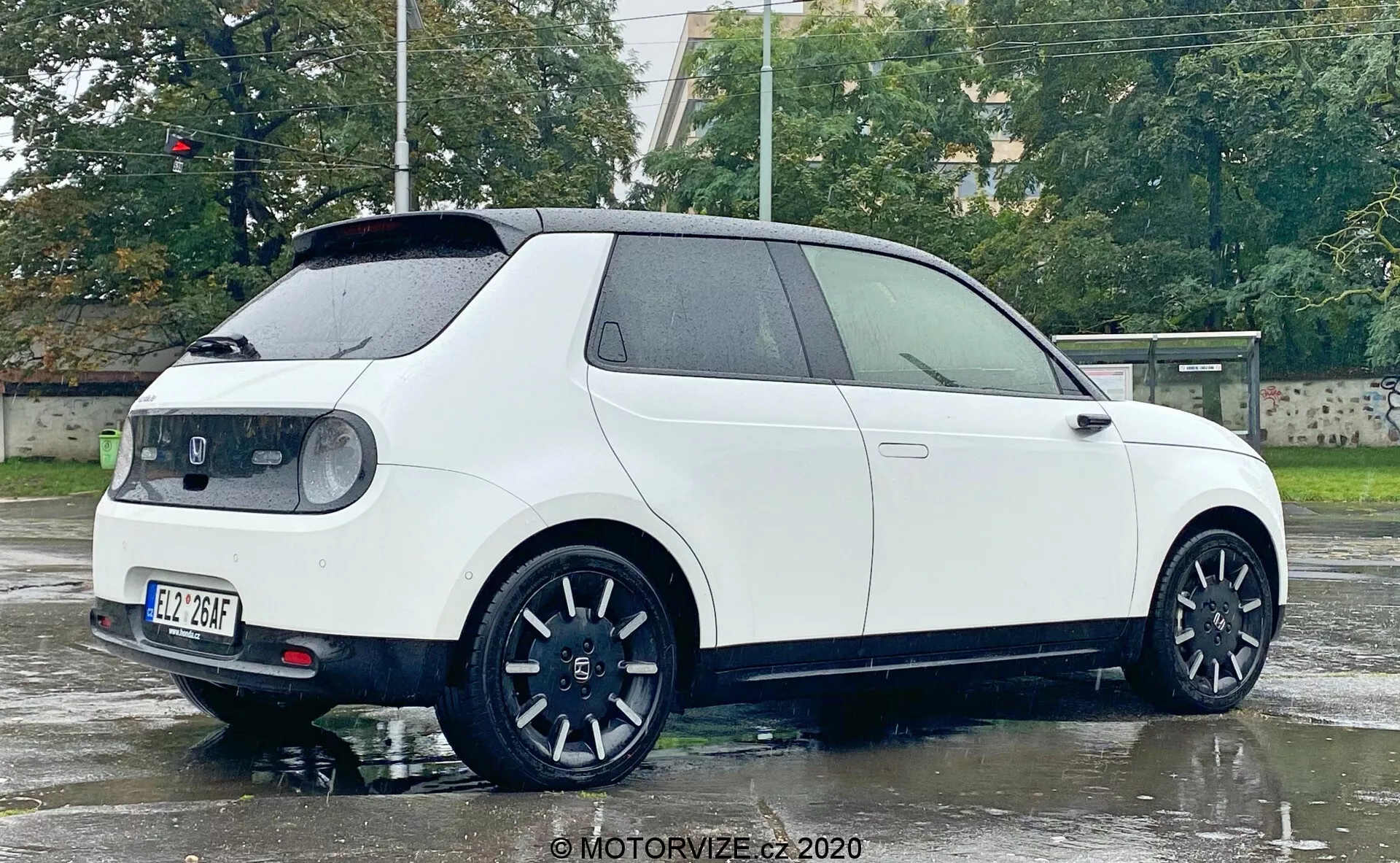 Obrázek ukazuje bok a zadní část bílého moderního hatchbacku Honda e 2020, pořízený z úhlu tří čtvrtin. Mokrá plocha a odrazy světla naznačují nedávný déšť. Viditelné prvky zahrnují boční a zadní profily, dvoje dveře, hranatý tvar s výraznými oblouky kol, velká černá kola se sportovním designem s více paprsky, malé zadní okno s stěračem, zadní střešní spoiler, výrazná zadní světla obepínající boky, centrálně umístěnou zadní státní poznávací značku a výjimečný odznak na zadních dveřích. Z technologických vlastností vyniká aerodynamika, sportovní design kol, moderní zadní světla pravděpodobně využívající LED technologii a zlepšená viditelnost zadního okna.