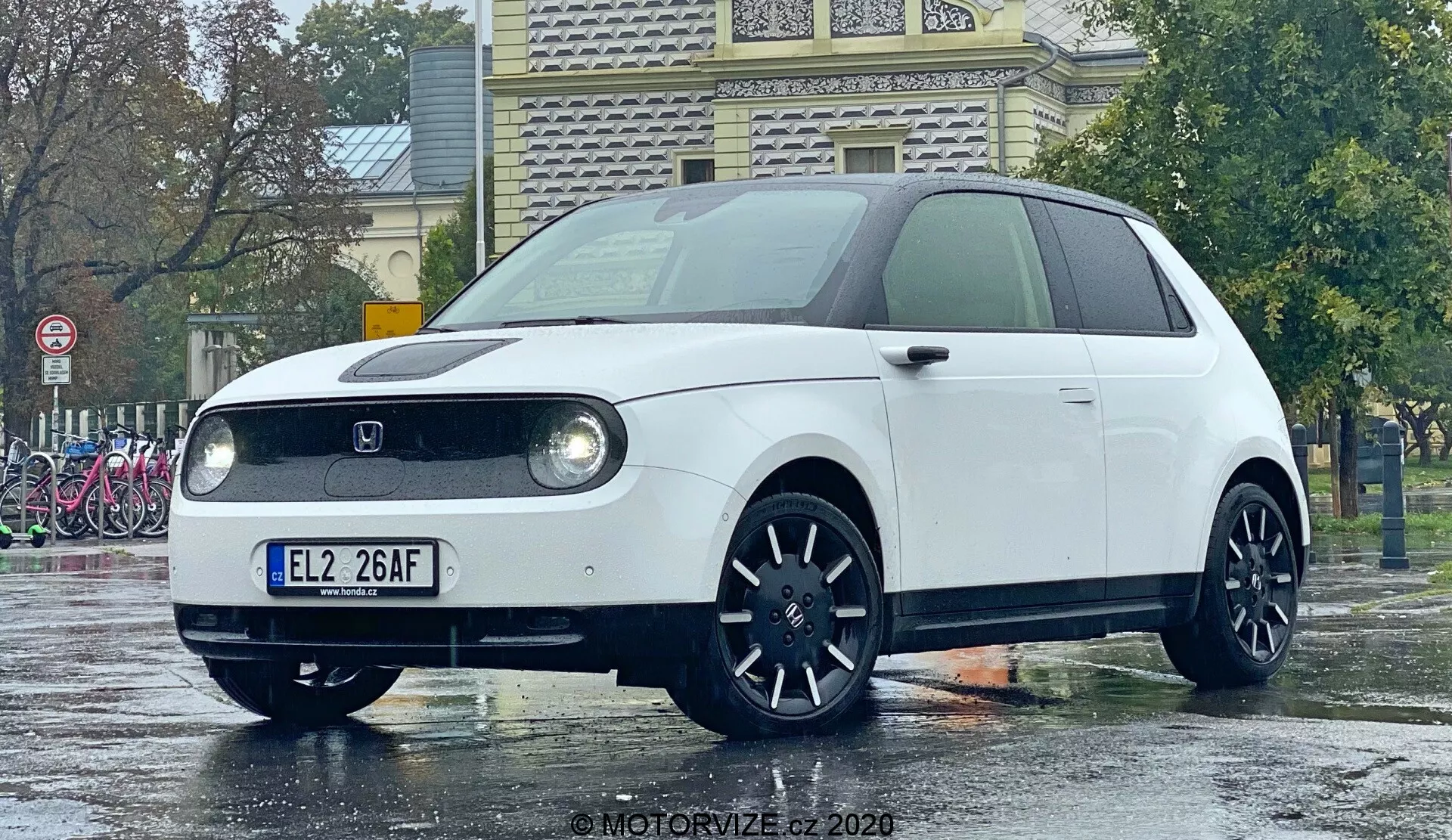 Honda e 2020 vyfotografovaná z nízkého úhlu zdůrazňujícího přední část a částečný boční profil, s kruhovými LED světlomety v obdélníkovém krytu, uzavřenou přední maskou typickou pro elektrická vozidla, centrálně umístěným logem výrobce, tmavými hliníkovými koly s mnoha paprsky a minimalistickým retro-moderním designem. Kompaktní hatchback stojí na mokré zemi odrážející deštivé prostředí, což zvýrazňuje jeho čisté linie a barvu.