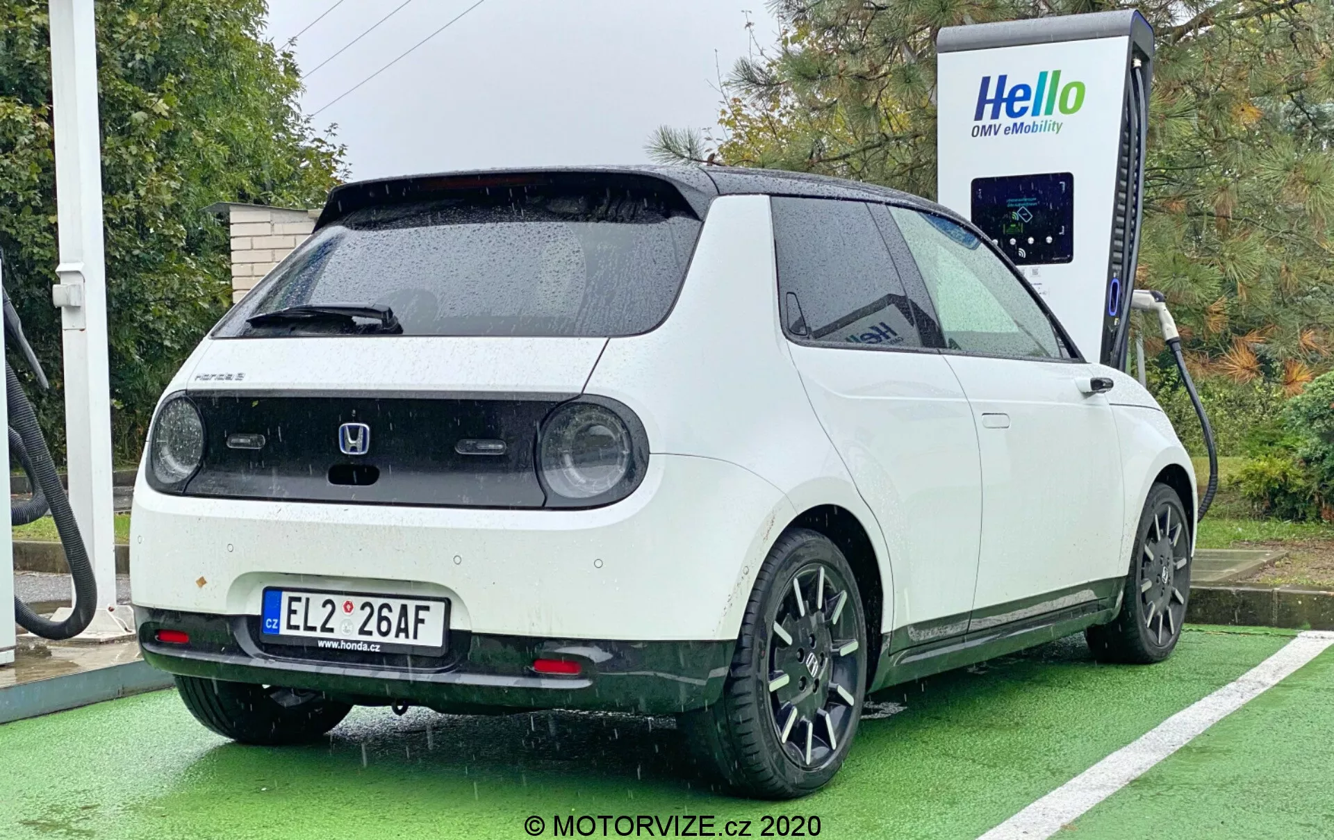 Elektrické vozidlo Honda e z roku 2020 je fotografováno z úhlu tři čtvrtiny zezadu, čímž zobrazuje pravý zadní čtvrtinový pohled, částečnou stranu a zadní dveře. Auto s dvoubarevným schématem, bílou karoserií a černou střechou, je zaparkované u nabíjecí stanice s připojeným nabíjecím kabelem na zadní straně. Design vozu zahrnuje kruhová zadní světla, velký zadní stěrač, parkovací senzory, střešní spoiler a černá pětipaprsková hliníková kola. Obrázek také zdůrazňuje hranatý, retro-moderní vzhled vozu, pořízený během dne za oblačného nebo deštivého počasí, s vozem zaparkovaným ve vyhrazeném místě pro nabíjení elektrických aut označeném zelenou barvou.