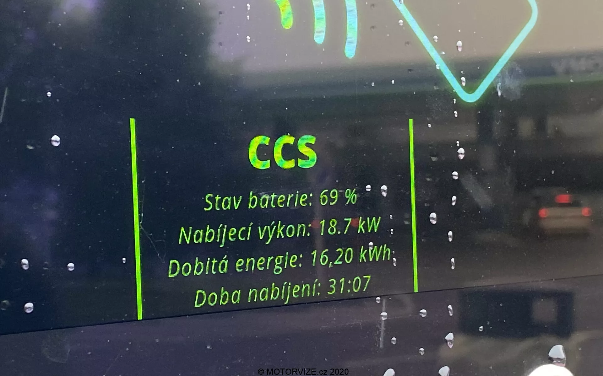 Pohled z interiéru elektrického vozidla přes čelní sklo, zaměřený na odraz rozhraní nabíjecí stanice na skle. Viditelné detaily zahrnují 'CCS' pro Combined Charging System, 'Stav baterie: 69 %,' 'Nabíjecí výkon: 18,7 kW,' 'Dobitá energie: 16,20 kWh,' a 'Doba nabíjení: 31:07.' Odraz působí duchovitě na deštivém pozadí s dalším vozidlem viditelným v dálce.