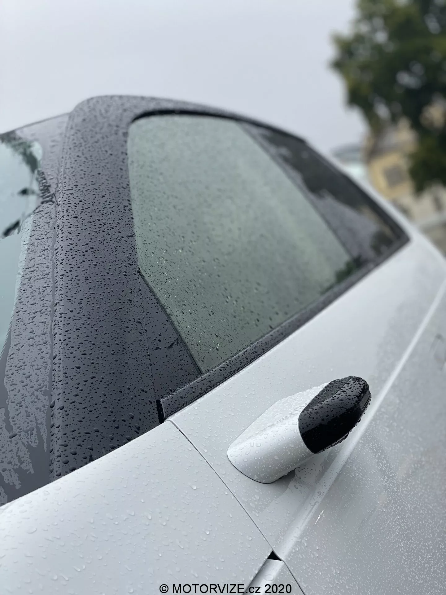 Detailní záběr na zadní část modelu Honda e 2020, zachycující konstrukci zadního bočního okna s kapkami vody a čisté světlé zadní dveře s černou vysouvací klikou dveří, zdůrazňující pokročilý nátěr a bezklíčkový vstupní systém.