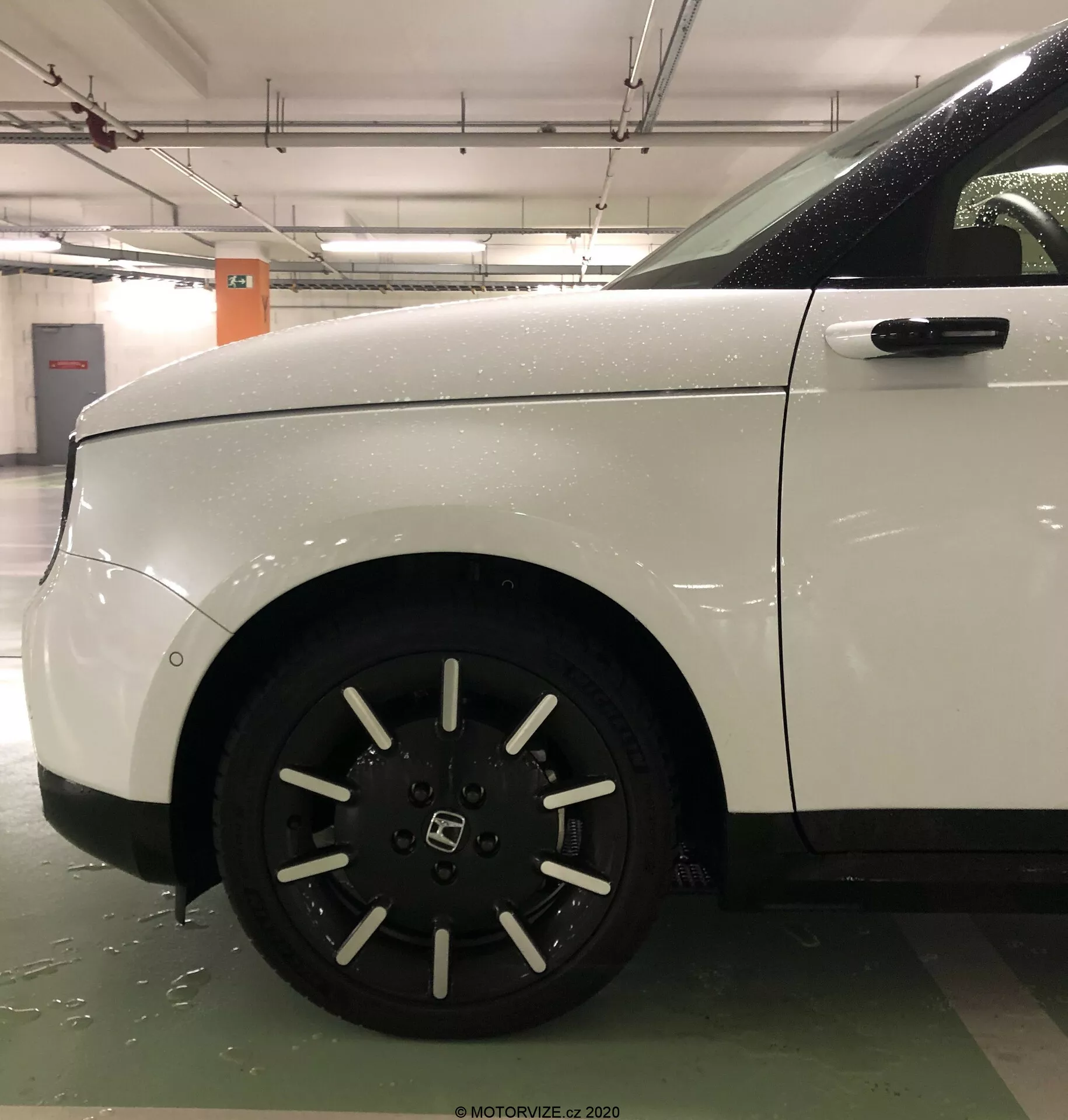 Detailní boční pohled na přední kolo a čtvrt panel bílého Honda E 2020, zobrazující desetipaprskové hliníkové kolo a čistou, aerodynamickou karoserii. Obraz, pravděpodobně pořízený v krytém parkovišti, zdůrazňuje udržovaný stav vozidla a robustní design, s viditelnými mokrými povrchy naznačujícími nedávné vystavení vodě.