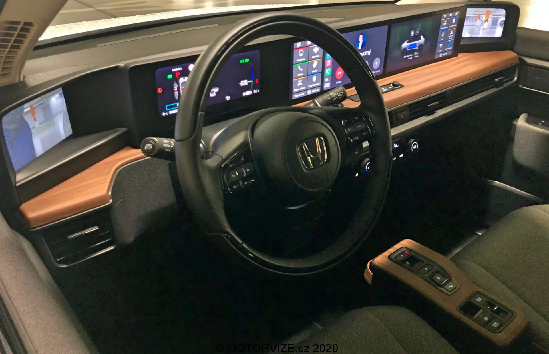Vnitřek vozu Honda e 2020 s pokročilými funkcemi, včetně multifunkčního volantu s ovládáním zvuku a hlasu, digitálního palubního displeje zobrazujícího rychlost a úroveň paliva, centrálního infotainment systému kompatibilního s Apple CarPlay a Android Auto, automatického dvouzónového klimatizačního systému, pohodlných kožených sedadel s funkcemi ohřevu, centrální konzole s řadicí pákou a úložištěm, pokročilých asistenčních systémů pro řidiče, možností připojení jako USB porty a bezdrátové nabíjení, prémiového zvukového systému a přizpůsobitelného ambientního osvětlení.