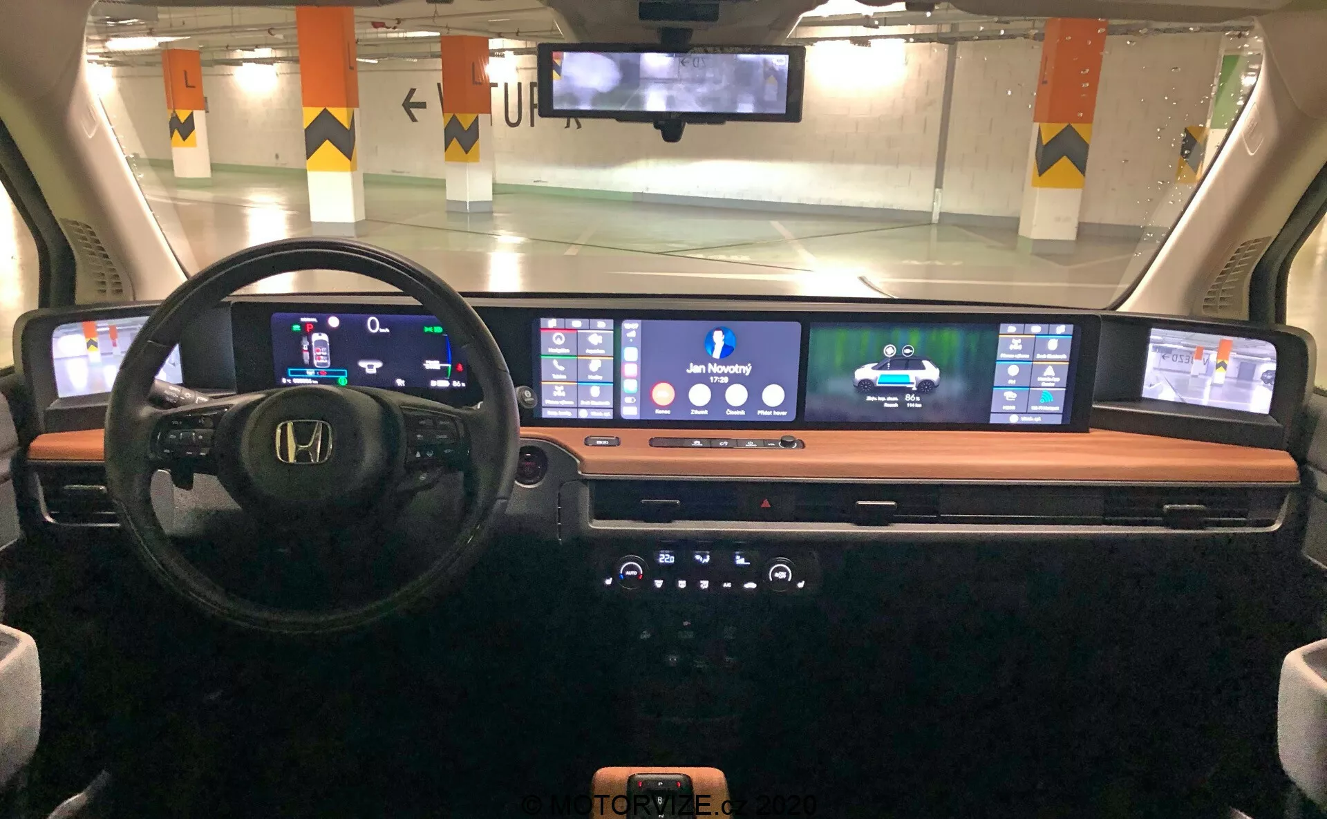 Interiérový pohled na palubní desku Honda e 2020 z pohledu řidiče, zobrazující digitální přístrojový panel, multifunkční volant, rozšířenou kontinuální digitální obrazovku pro navigaci a média, elegantní středovou konzoli s knoflíky a tlačítky a bezdrátovou nabíjecí podložku, zvýrazňující moderní automobilovou technologii a design.