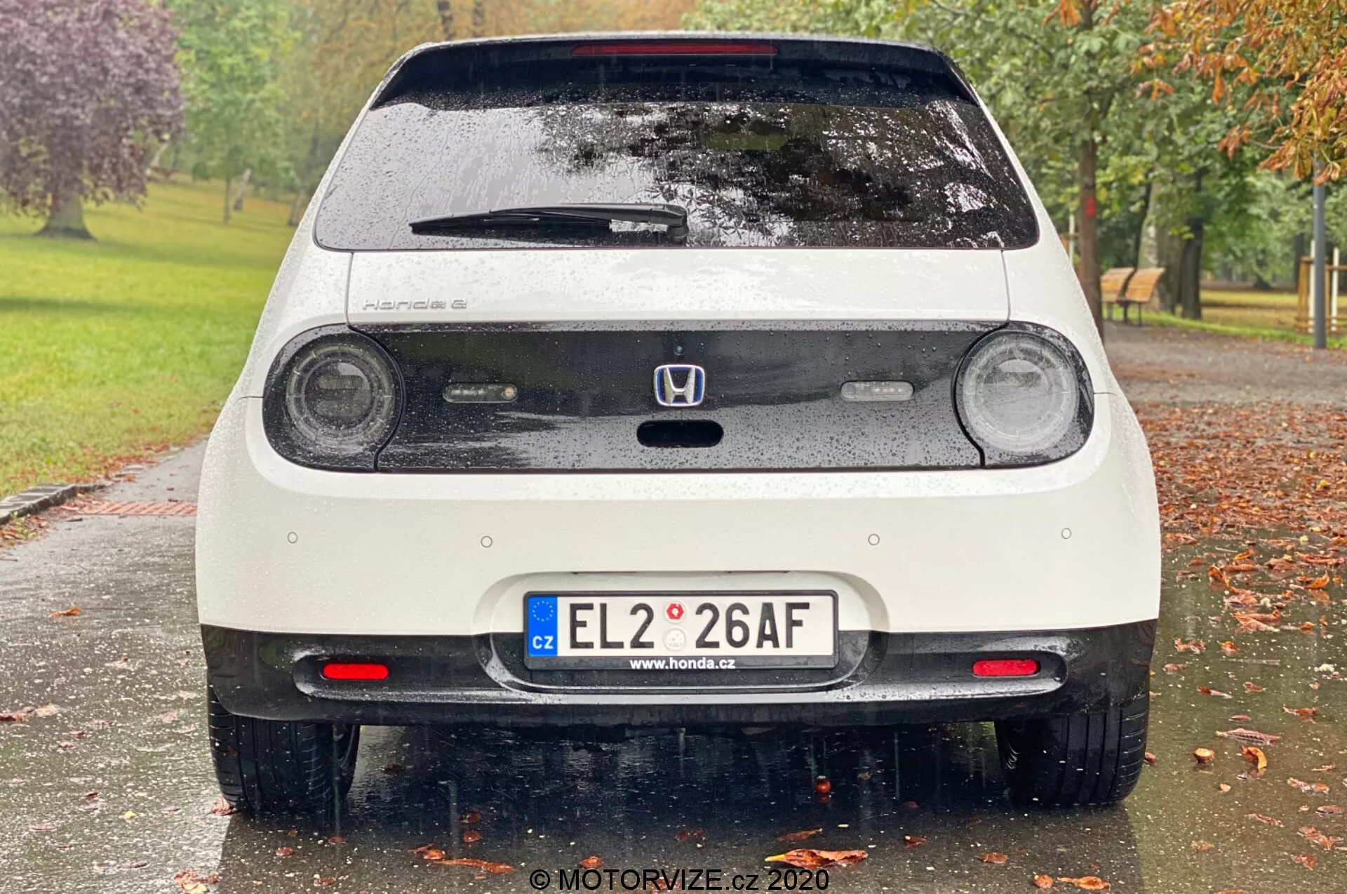 Zadní pohled na mokré bílé auto Honda e 2020, které zobrazuje kruhová LED zadní světla, třetí brzdové světlo, emblém Honda, zadní dveře s stěračem okna, evropskou SPZ, parkovací senzory, zadní mlhová světla, zadní spoiler a reflexní prvky na nárazníku. Obrázek zachycuje jemné detaily jako kapky vody na vozidle, s rozmazaným parkovým pozadím s stromy a trávou.