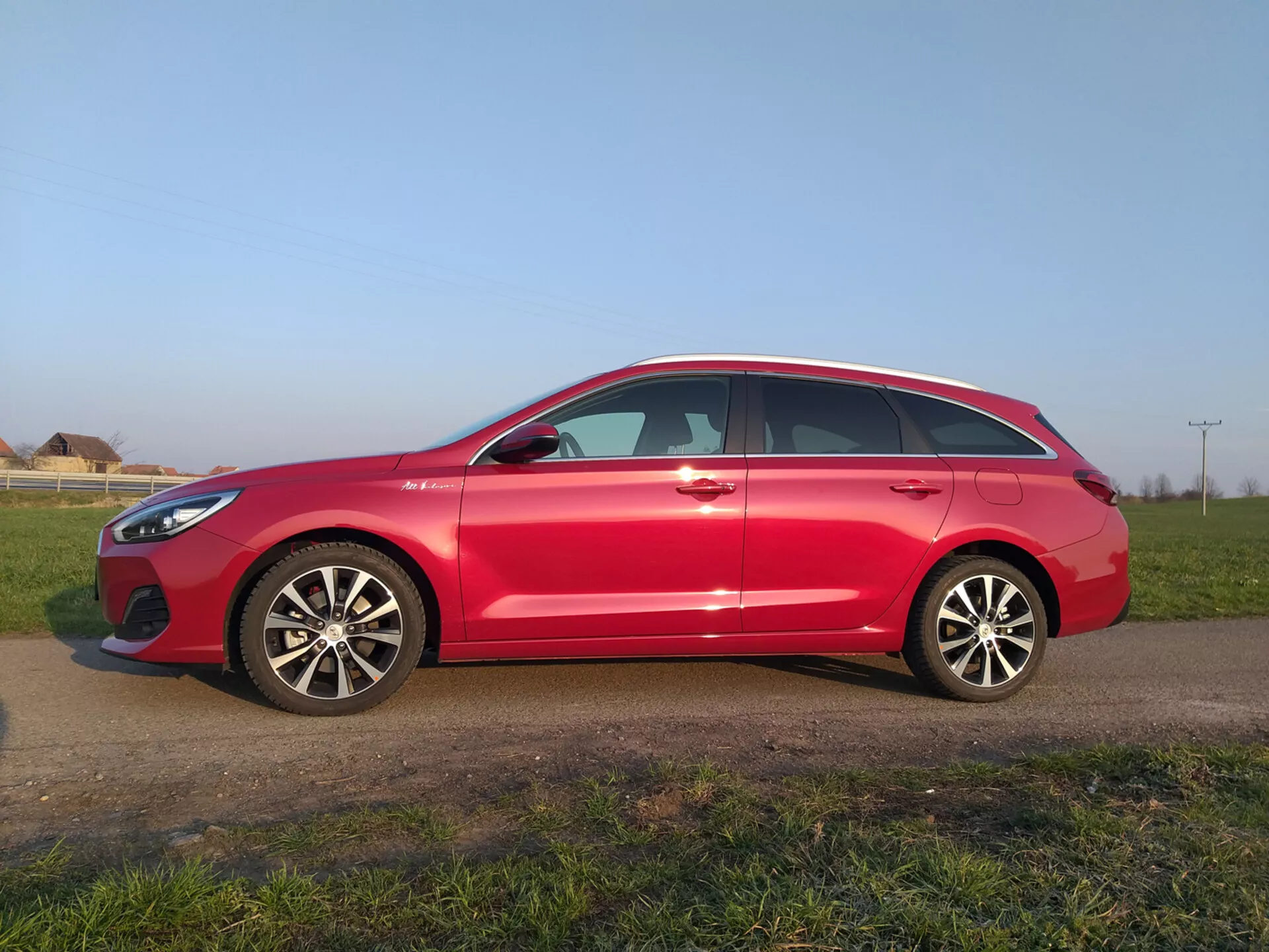 Červený Hyundai i30 Fastback 2017 s moderním aerodynamickým profilem se svažující se střechou, slitinovými koly s více paprsky, zrcátky se zabudovanými směrovými signály, okny s chromovým rámem a pneumatikami připravenými na silnici, zaparkovaný na travnatém poli pod jasným nebem.