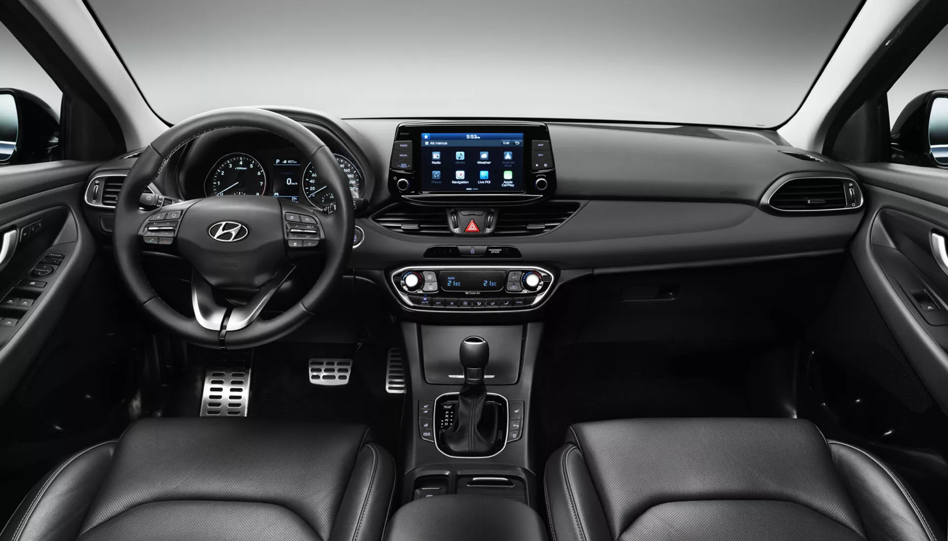 Interiér Hyundai i30 2017 Fastback zobrazující přední palubní desku z perspektivy zadního sedadla, s volantem umístěným uprostřed vlevo s tlačítky ovládání a logem Hyundai, digitálním přístrojovým panelem, velkým dotykovým infotainment systémem s aplikacemi pro připojení, duálním klimatizací, pákou automatické převodovky, potenciálními ovládacími prvky pro vytápění/chlazení sedadel a sedadla čalouněná kůží.