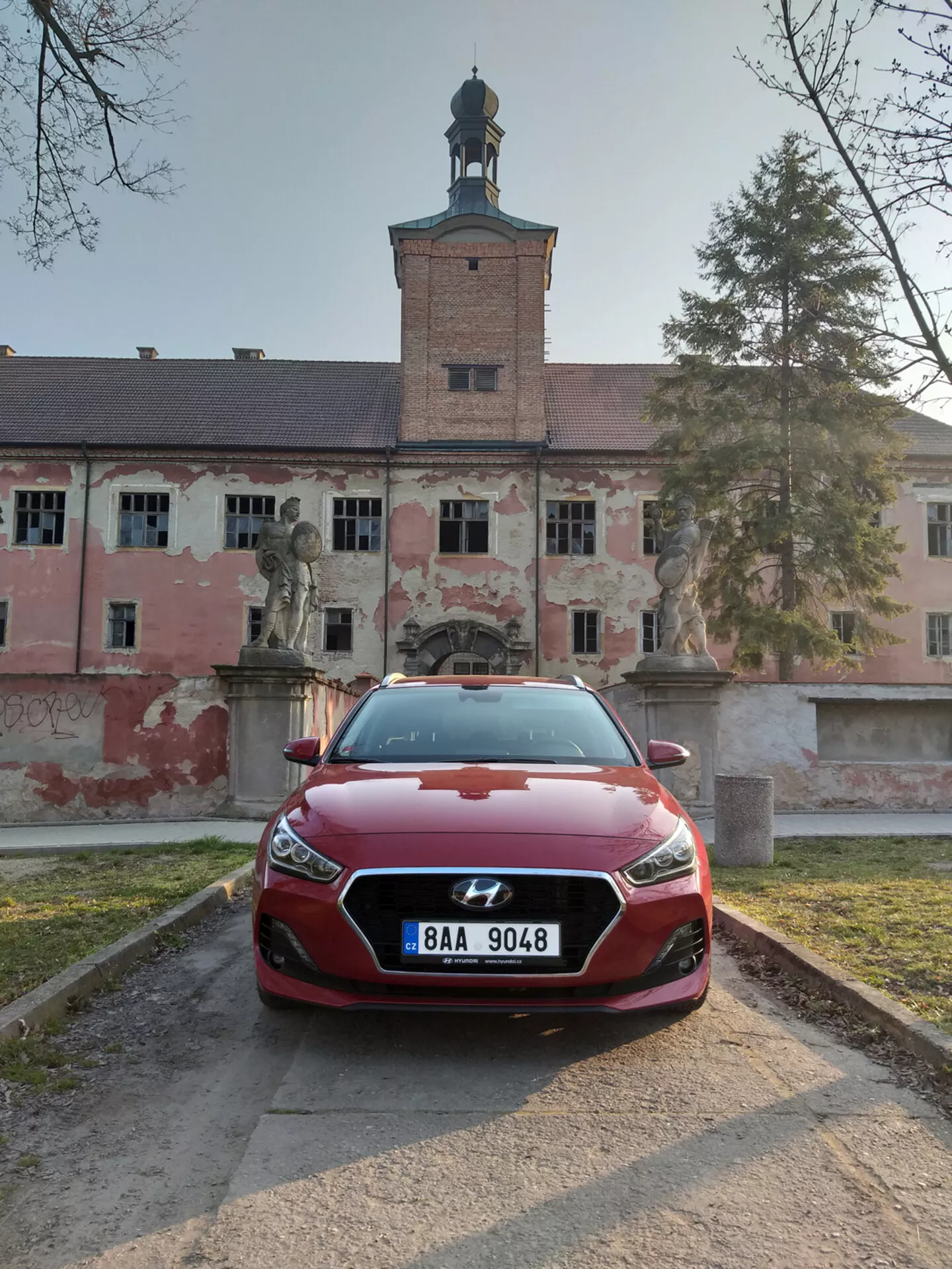 Červený fastback Hyundai i30 2017 s pohledem zepředu na LED světlomety a výrazný design mřížky, umístěný na štěrkové cestě s historickou budovou v pozadí.
