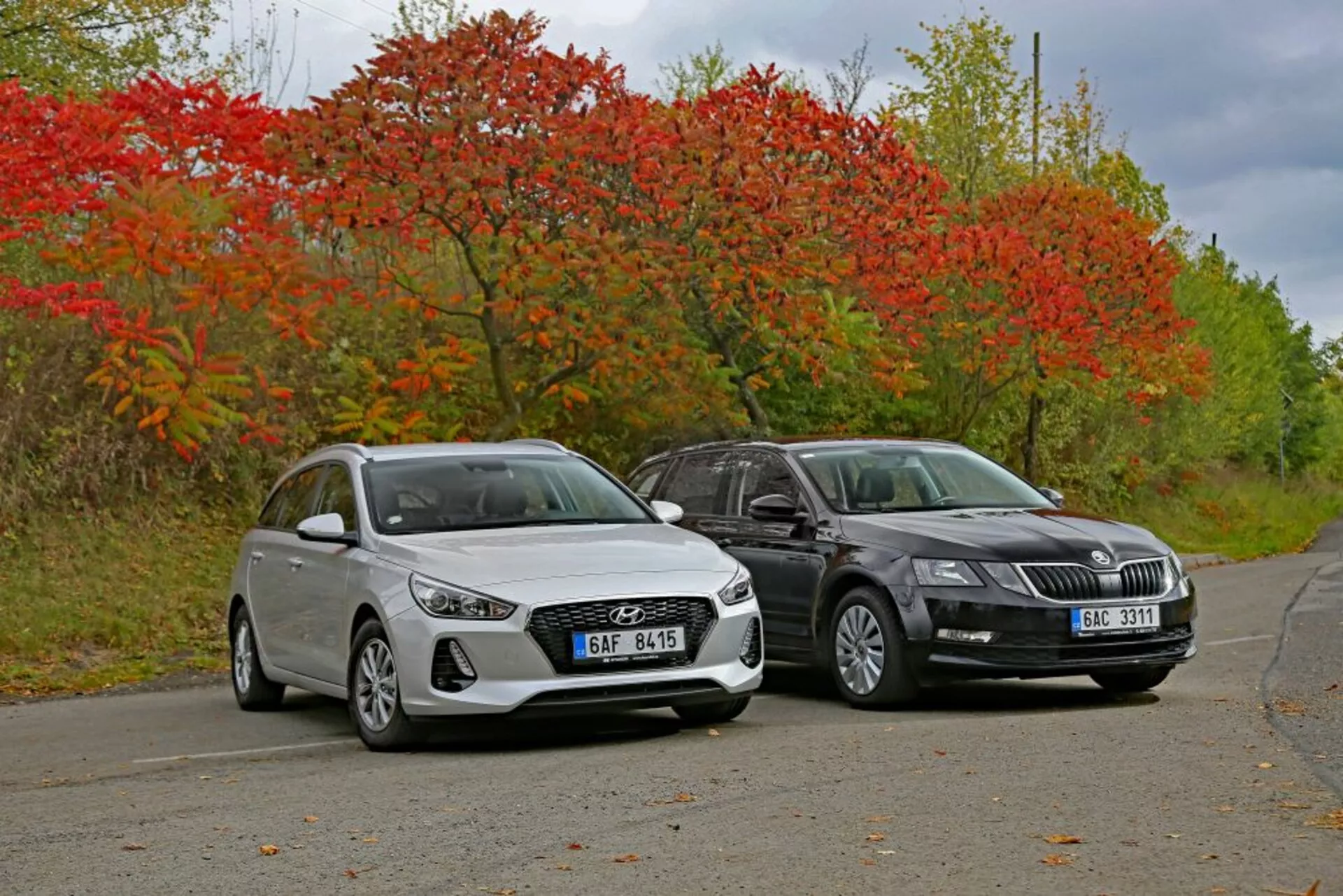 Bílý hatchback Hyundai i30 z roku 2017 a tmavě zbarvený sedan vedle sebe na klidné silnici obklopené podzimní vegetací, předvádějící designové linie a moderní osvětlovací technologie