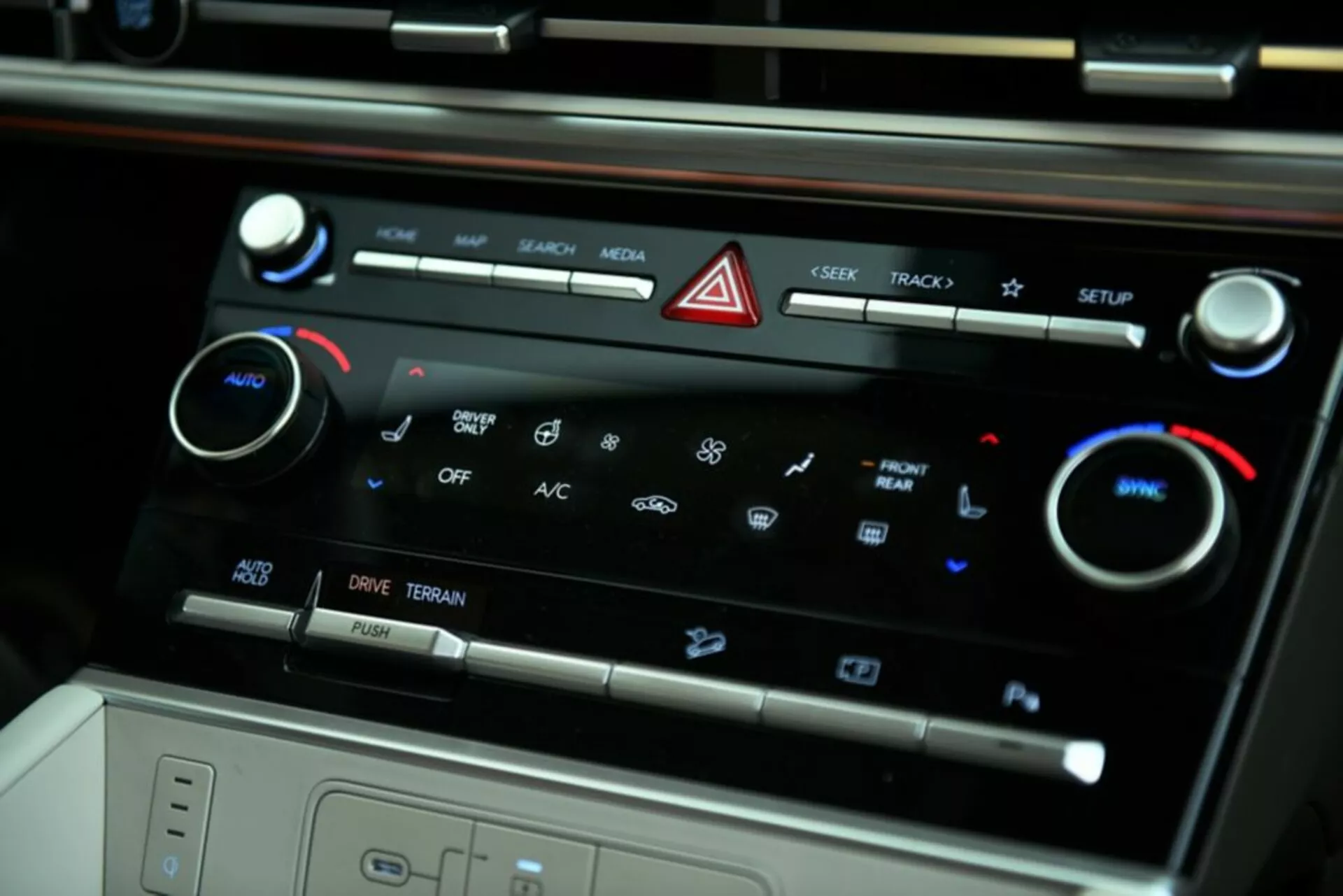 Blízký pohled z úhlu na centrální ovládací panel klimatizace modelu Hyundai Santa Fe 2024. Obrázek zvýrazňuje dotyková tlačítka pro ovládání médií, tlačítko nouzových světel, dvouzónovou klimatizaci se zobrazenými ikonami, nastavovací délky teploty, aktivaci A/C, ovládání rychlosti ventilátoru, možnosti jízdních režimů a náznak úložného prostoru nebo bezdrátové nabíjecí podložky.