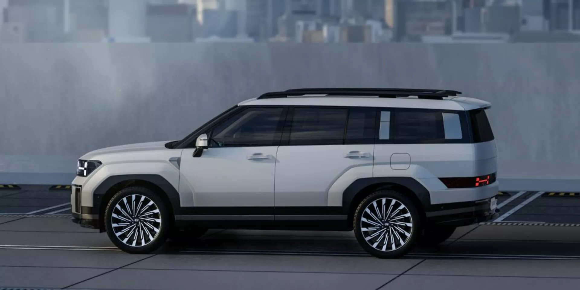 Boční profil SUV Hyundai Santa Fe z roku 2024, který předvádí svůj hranatý, ale moderní design s výraznými liniemi a hranami. Viditelné prvky zahrnují boční dveře, okna, boční zrcátka, velká vícepaprsková hliníková kola, dvoubarevný lak s světlým tělem a tmavší střechou a spodním lemem, vysokou světlou výšku, stupačky a střešní ližiny. Na pozadí je neurčité rozmazané městské prostředí.