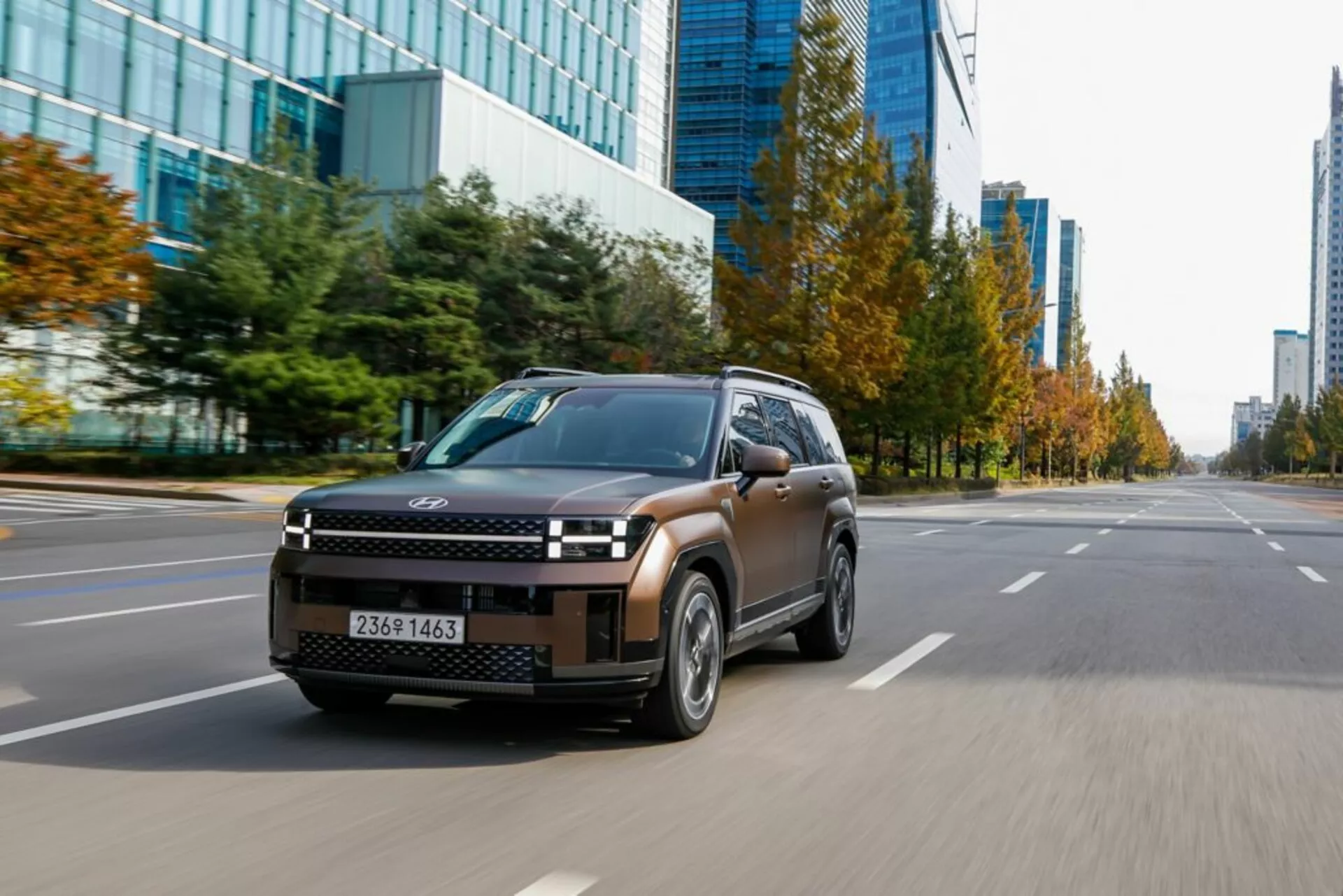 Obrázek modelu Hyundai Santa Fe 2024 páté generace, pohled z tři čtvrtiny zepředu zleva, pořízený z nízkého úhlu ukazujícího elegantní přední masku SUV, LED světlomety, kapotu, přední blatníky, čelní sklo, boční dveře a boční zrcátka. Auto se pohybuje po městské silnici s několika pruhy a komerčními budovami v pozadí. Aerodynamická kola a částečně rozmazaná poznávací značka zdůrazňují pohyb a výkon vozidla v městském prostředí.