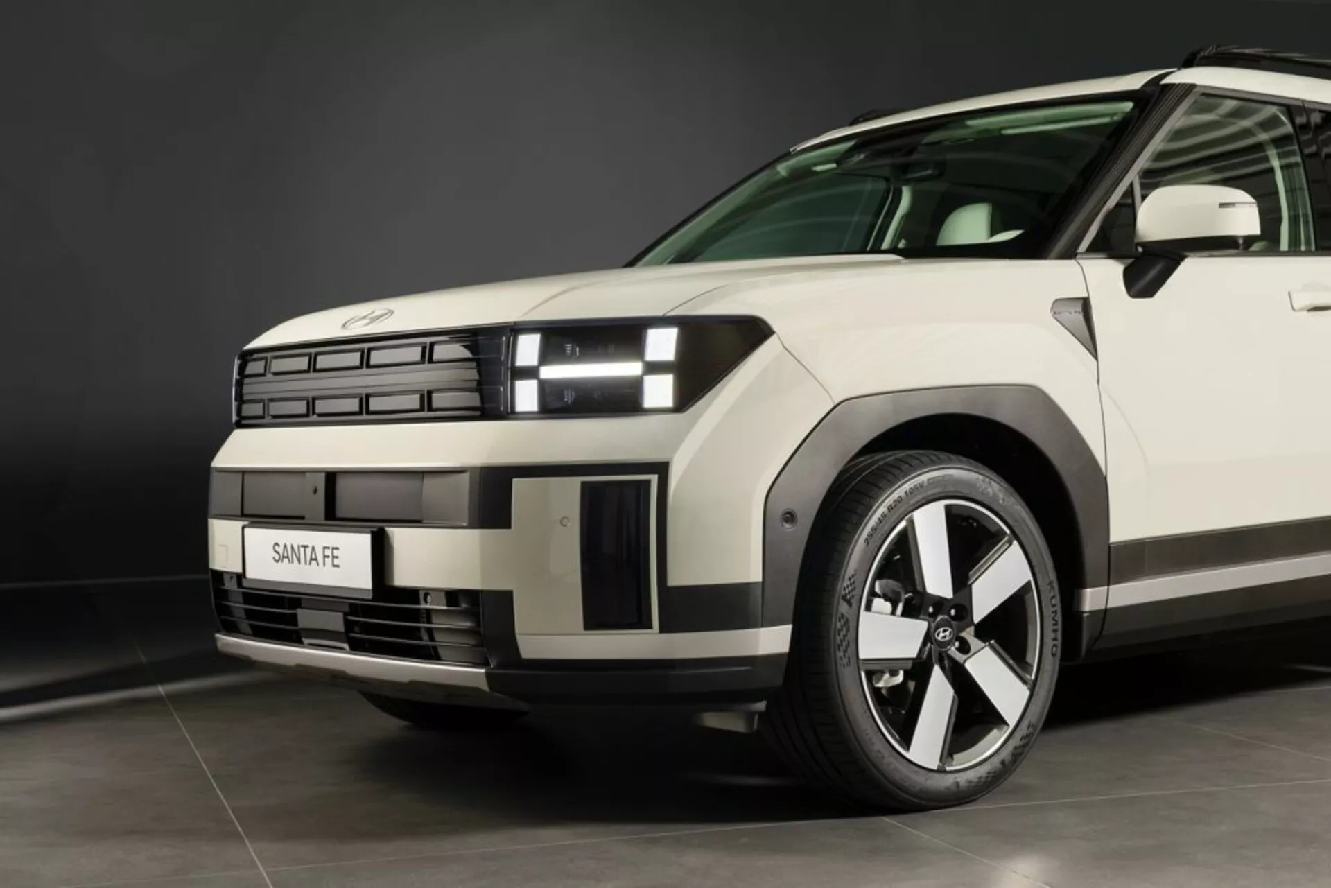 Blízký pohled z nízkého úhlu na Hyundai Santa Fe 2024 (5. generace) s důrazem na přední levý čtvrtý díl, ukazující moderní design přední masky, dělené světlomety se specifickým denním svícením, sochanou kapotu, víceúčelové litinové kolo, viditelný běhoun pneu, značkovou masku, zřetelnou linii na blatníku, aerodynamický přední levý dveřní panel a zrcátko, a neutrální barvu pod studiovým osvětlením.