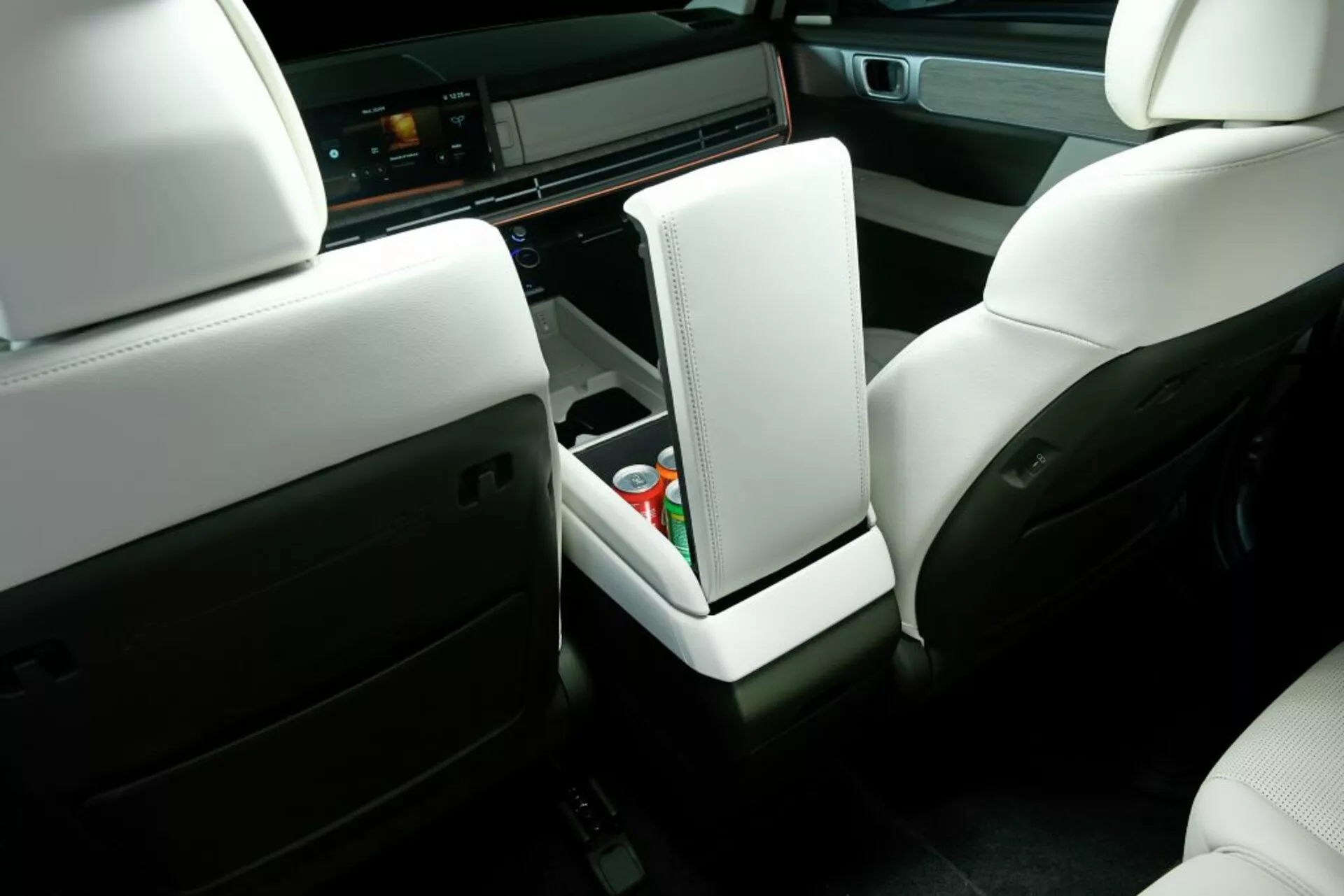 Pohled na interiér z perspektivy zadního sedadla vozu Hyundai Santa Fe 2024 (5. generace), zobrazující světlé kožené přední sedadla s opěrkami hlavy, vzpřímenou centrální opěrku, středovou konzoli s otevřeným úložným prostorem obsahujícím plechovku s nápojem a drobné předměty, moderní infotainment systém na palubní desce zobrazující rozhraní a směs světlých a tmavých materiálů zdůrazňující prémiový a komfortní design.
