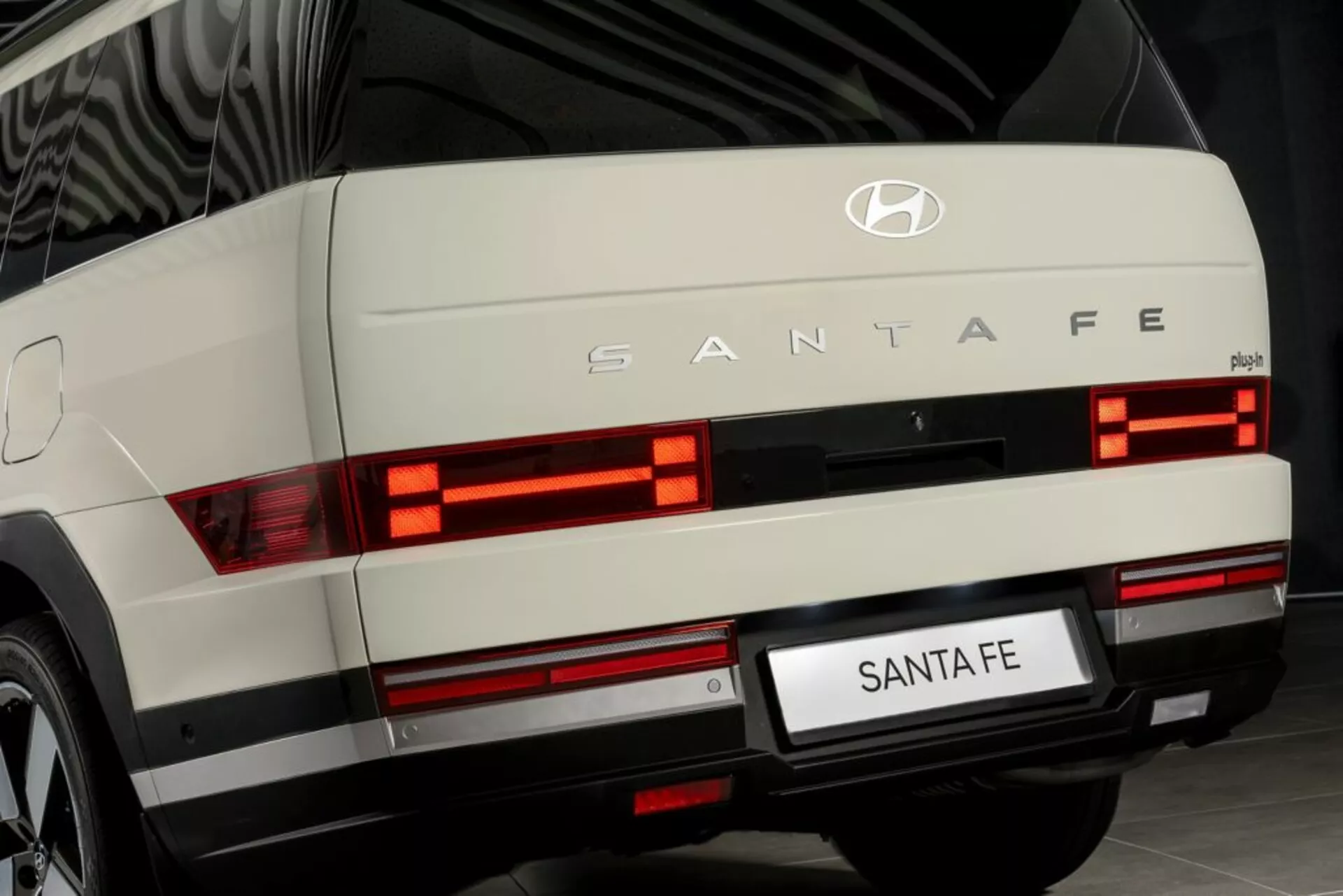 Blízký pohled na zadní část Hyundai Santa Fe 2024 (5. generace), pořízený z úrovně země. Obrázek ukazuje osvětlené vodorovné, segmentované zadní světla, stříbrný ozdobný prvek, znak Hyundai a označení 'SANTA FE'. Viditelné jsou zadní stěrač, oblast zadní registrační značky a možná mlhová světla nebo odrazky na úrovni nárazníku. Zobrazena je v interiéru, což naznačuje dobře udržované moderní vozidlo možná použití pro propagační účely.