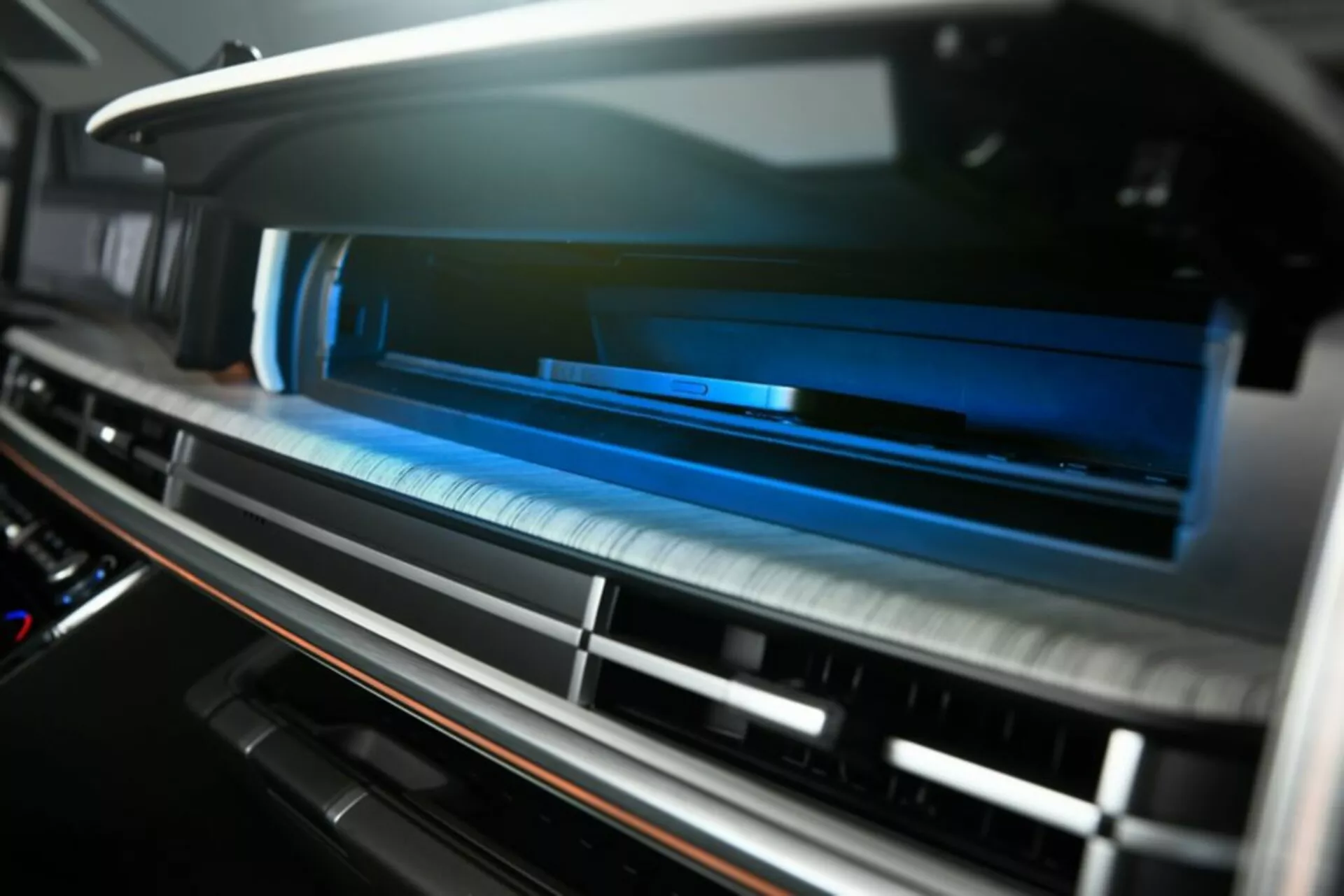 Detailní záběr na větrací systém uvnitř palubní desky Hyundai Santa Fe 2024 5. generace, pohled mírně zespodu. Horizontální lamely ventilu v téměř uzavřené poloze se sníženou hloubkou ostrosti, rozmazání popředí a pozadí. Moderní kartáčovaný kovový povrch a potenciální dotykově citlivé ovládání, zvýrazněné modrým ambientním osvětlením pro high-tech, prémiový interiér. Kontrastní materiálový okraj naznačuje luxusní povrchovou úpravu, možná dřevo nebo imitace dřeva. Žádné viditelné knoflíky/tlačítka naznačují moderní dotykový systém HVAC. Snímek interiéru se zaměřuje na pohodlí, pohodlí a pokročilé funkce klimatizace.
