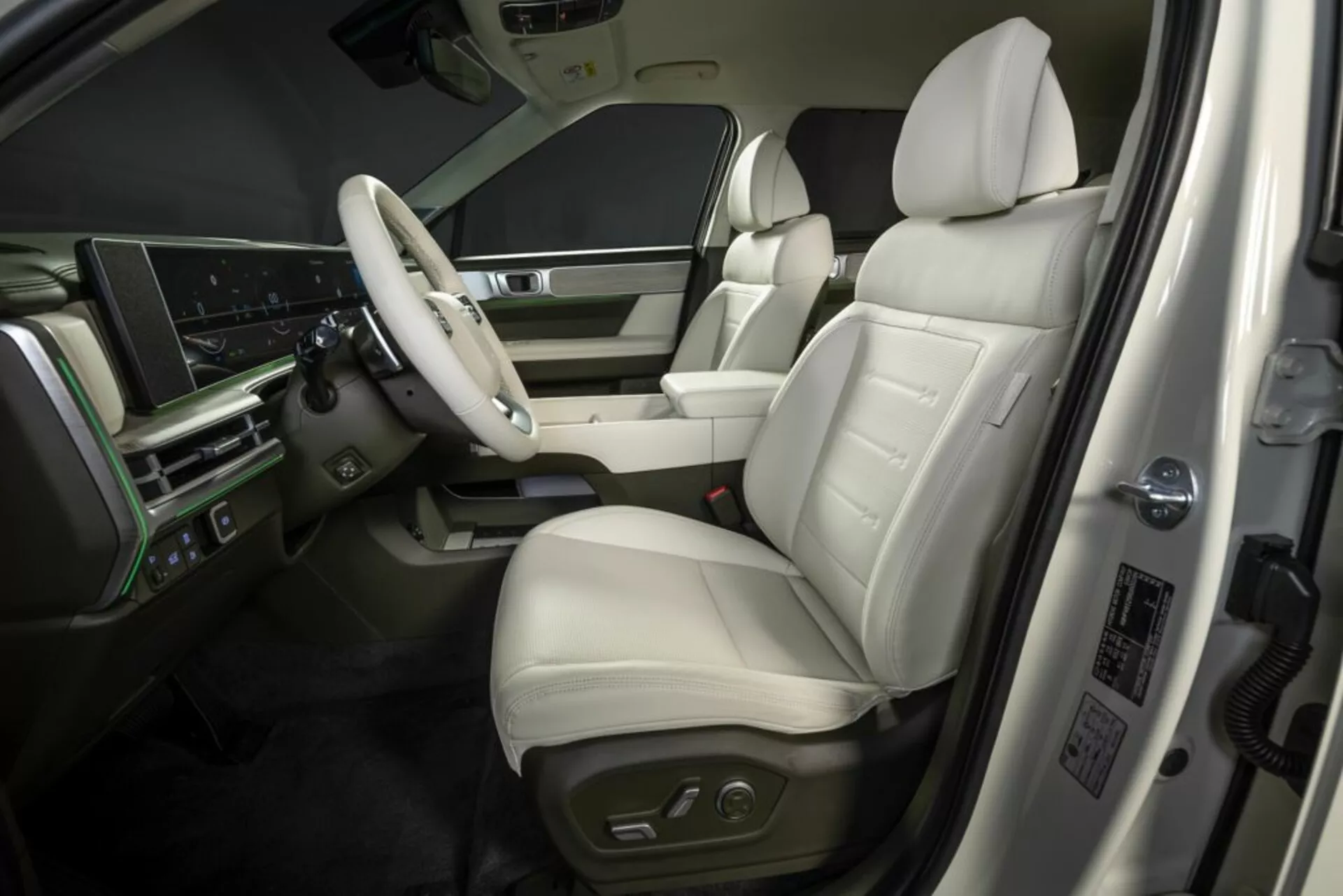 Obrázek ukazuje luxusní interiér Hyundai Santa Fe 2024 (5. generace) z vyvýšeného vnějšího úhlu. Zaměřuje se na oblast přední strany řidiče, zahrnuje kožené sedadlo s elektronickým nastavením, panel dveří s ovládáním oken a zrcátek, multifunkční volant s funkcemi infotainmentu a tempomatu, velký digitální displej přístrojové desky a detaily z imitace dřeva. Středová konzola naznačuje další ovládací prvky a řadicí páku automatické převodovky, zatímco na dveřním sloupku jsou zobrazeny údaje o výrobě nebo bezpečnostní informace.