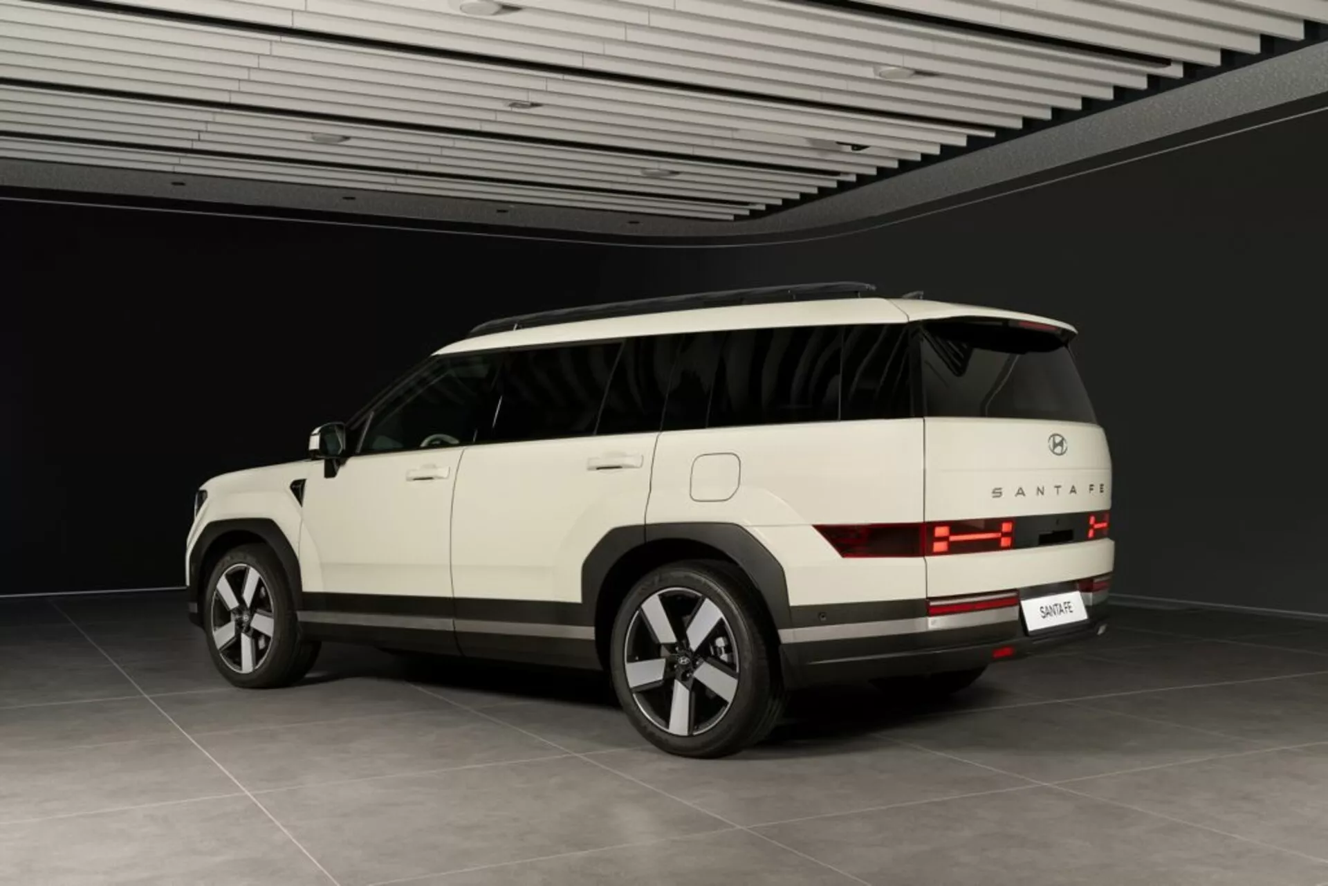 Tříčtvrteční zadní pohled na SUV Hyundai Santa Fe 2024 (5. generace) v neutrálním vnitřním prostředí. Viditelné jsou zadní světla s horizontálními čarami, zadní okno, nárazník a prostor pro poznávací značku. Elegantní boční profil ukazuje kliky dveří, obrysové linie a částečně viditelnou panoramatickou střechu. Moderní prvky zahrnují velká slitinová kola s více paprsky, boční zrcátko s integrovanými směrovými světly a tónovaná skla. Světle zbarvené SUV a kontrolované prostředí naznačují, že jde o luxusní model s pokročilou technologií.
