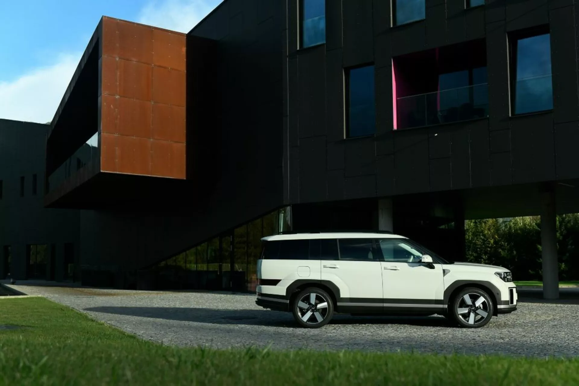 Boční profil Hyundai Santa Fe 2024 (5. generace) v bílé barvě, zaparkovaného před moderní černou budovou s oranžovou částí. Obrázek ukazuje linie karoserie SUV, boční dveře, okna, boční zrcátka a vícepaprsková litá kola. Kompozice zdůrazňuje kontrast mezi moderním designem vozidla a současnou architekturou.