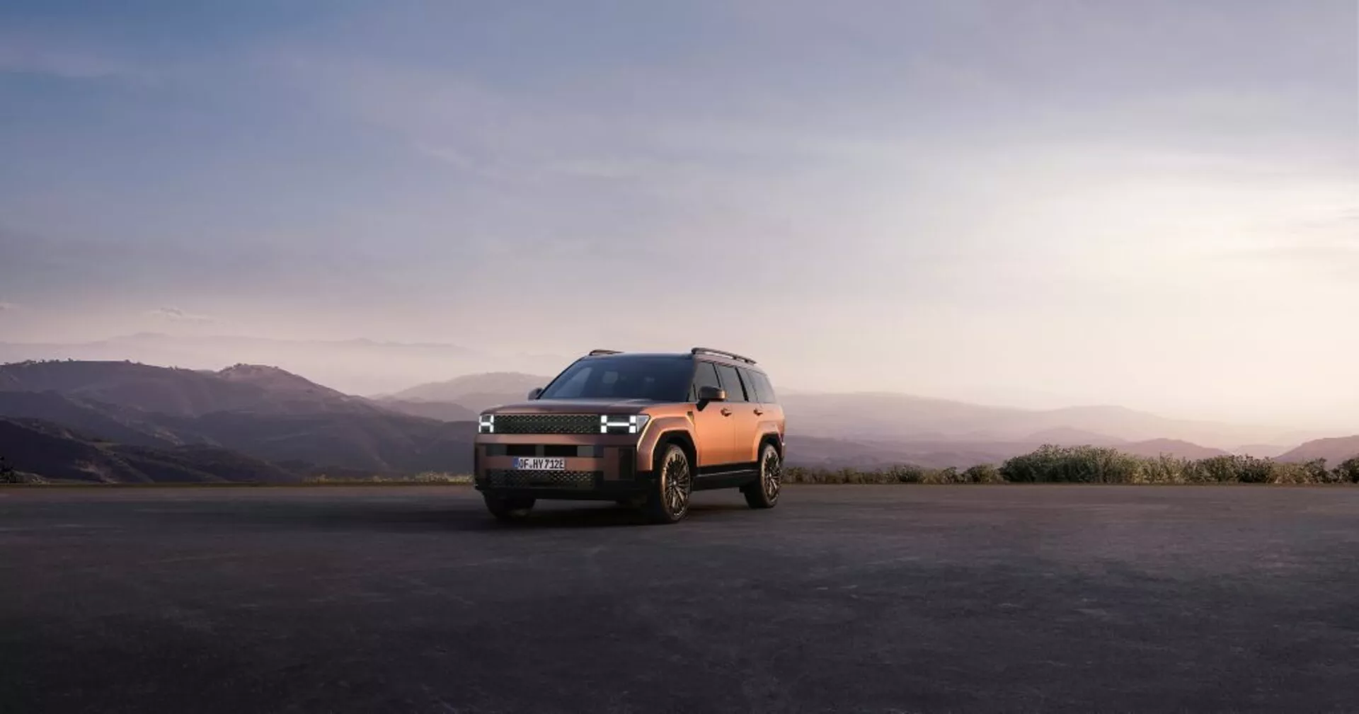 Pohled z čtvrtiny čela modelu Hyundai Santa Fe 2024 (5. generace) SUV zachyceného venku na asfaltovém povrchu, vyznačující se elegantním a zároveň drsným designem s LED světlomety, výraznou přední mřížkou a dvoubarevným tělem s tmavým ochranným obložením. Snímek byl pořízen během východu nebo západu slunce s kopcovitou krajinou v rozmazaném pozadí, zvýrazňující moderní estetiku vozidla a možný elektrický nebo hybridní pohonný systém.