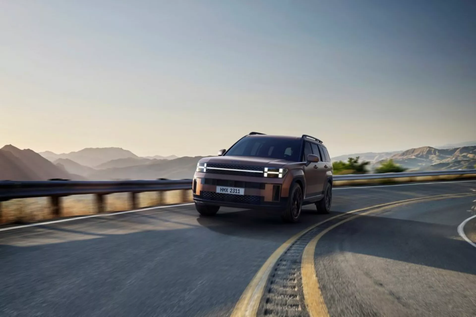 SUV Hyundai Santa Fe 2024 (5. generace) zachycený v dynamické perspektivě zepředu ze tří čtvrtin na klikaté silnici, zvýrazňující jeho moderní a elegantní design. Výrazně je zobrazena levá strana vozidla, ukazující charakteristická LED denní světla rámující odvážnou přední masku, úzké světlomety a víceramenná litá kola. Vozidlo je v pohybu, což demonstruje jeho robustní terénní schopnosti a pokročilé technologické prvky jako ADAS a adaptivní světlomety, s matným, vysoce kvalitním povrchem.