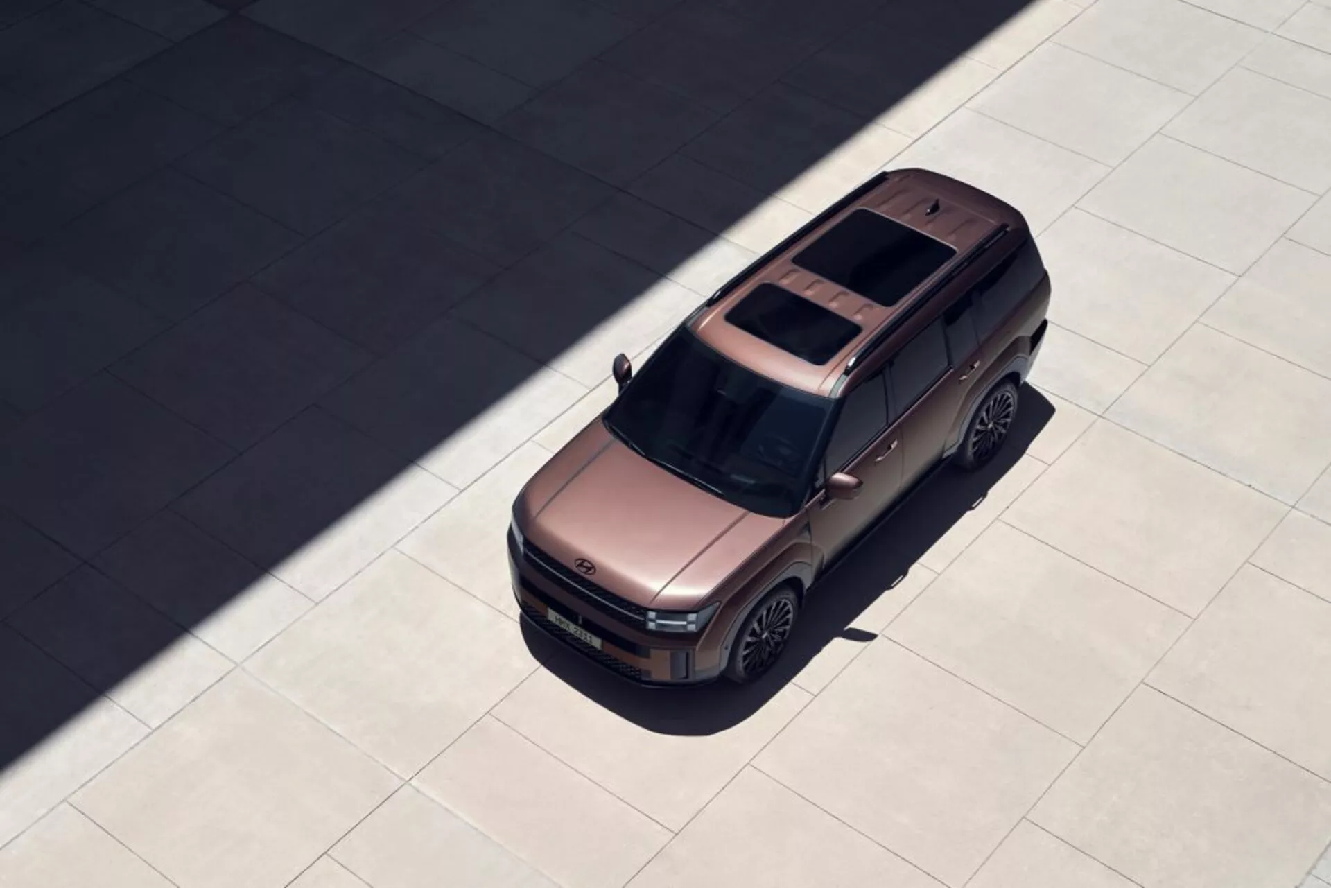 Fotografie Hyundai Santa Fe 2024 (5. generace) z vrchního úhlu, která zdůrazňuje jeho hranatý, robustní design, panoramatické střešní okno, zadní anténu, konturovanou kapotu, složená boční zrcátka, velká slitá kola a stín naznačující vysoce kontrastní osvětlení. SUV je zaparkováno na hladkém povrchu, což nabízí jasný pohled na jeho moderní a vizuálně atraktivní vlastnosti.
