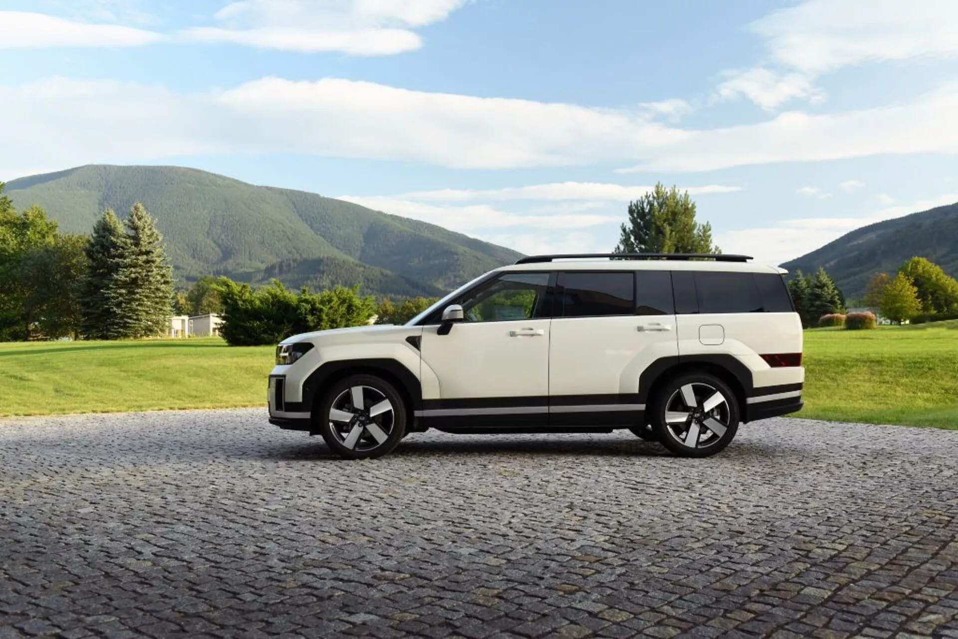 Boční pohled na SUV Hyundai Santa Fe (5. generace) z roku 2024 zaparkované na dlážděném povrchu s přírodním pozadím. Vozidlo má dvoubarevnou karoserii s bílou barvou a černou střechou, velká hliníková kola, žádné viditelné kliky dveří, což naznačuje zatahovací technologii, velká okna, střešní nosiče a vysokou světlou výšku. Ideální pro představení automobilového designu, luxusu, technologických vylepšení a schopností v terénu.
