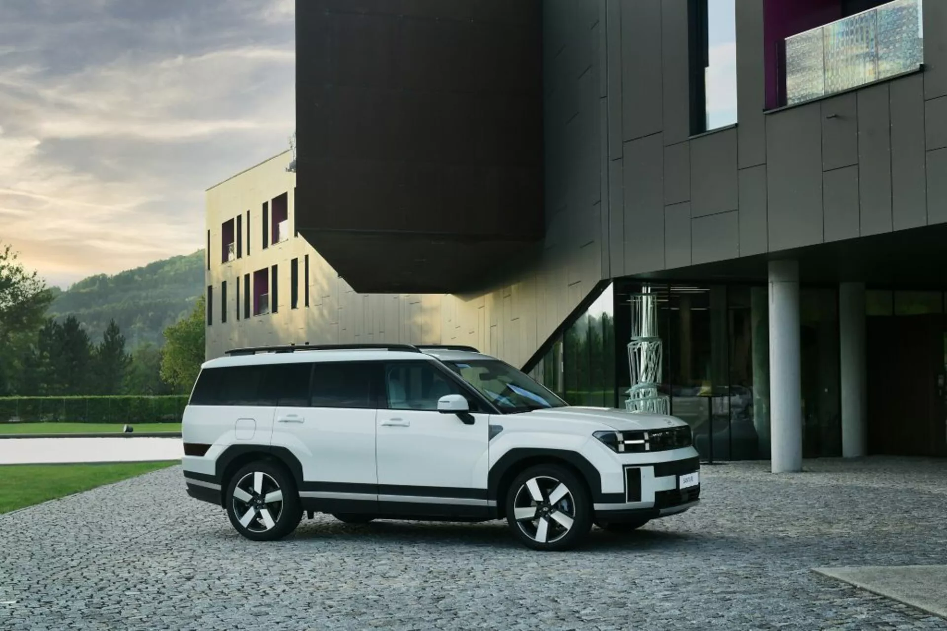 Na obrázku je moderní bílý Hyundai Santa Fe 2024 SUV (5. generace) zaparkovaný na dlážděném příjezdovém cestě před moderní budovou. Záběr z tříčtvrteční čelní perspektivy ukazuje SUV s elegantní přední mřížkou integrovanou s horizontálními světlomety, černě lemovaným nárazníkem a mlhovkami. Boční profil odhaluje velká okna s černými obrysy, zapuštěné kliky dveří a silnou ramenní linii. Vícepaprsková, dvoubarevná kola doplňují design vozidla, zatímco plochá střecha s potenciálním panoramatickým střešním oknem a zadním spoilerem zvyšuje jeho aerodynamický styl. Obrázky naznačují LED technologii v osvětlovacím systému, což zvýrazňuje moderní estetiku vozidla. Současná architektura pozadí přispívá k luxusní prezentaci SUV.