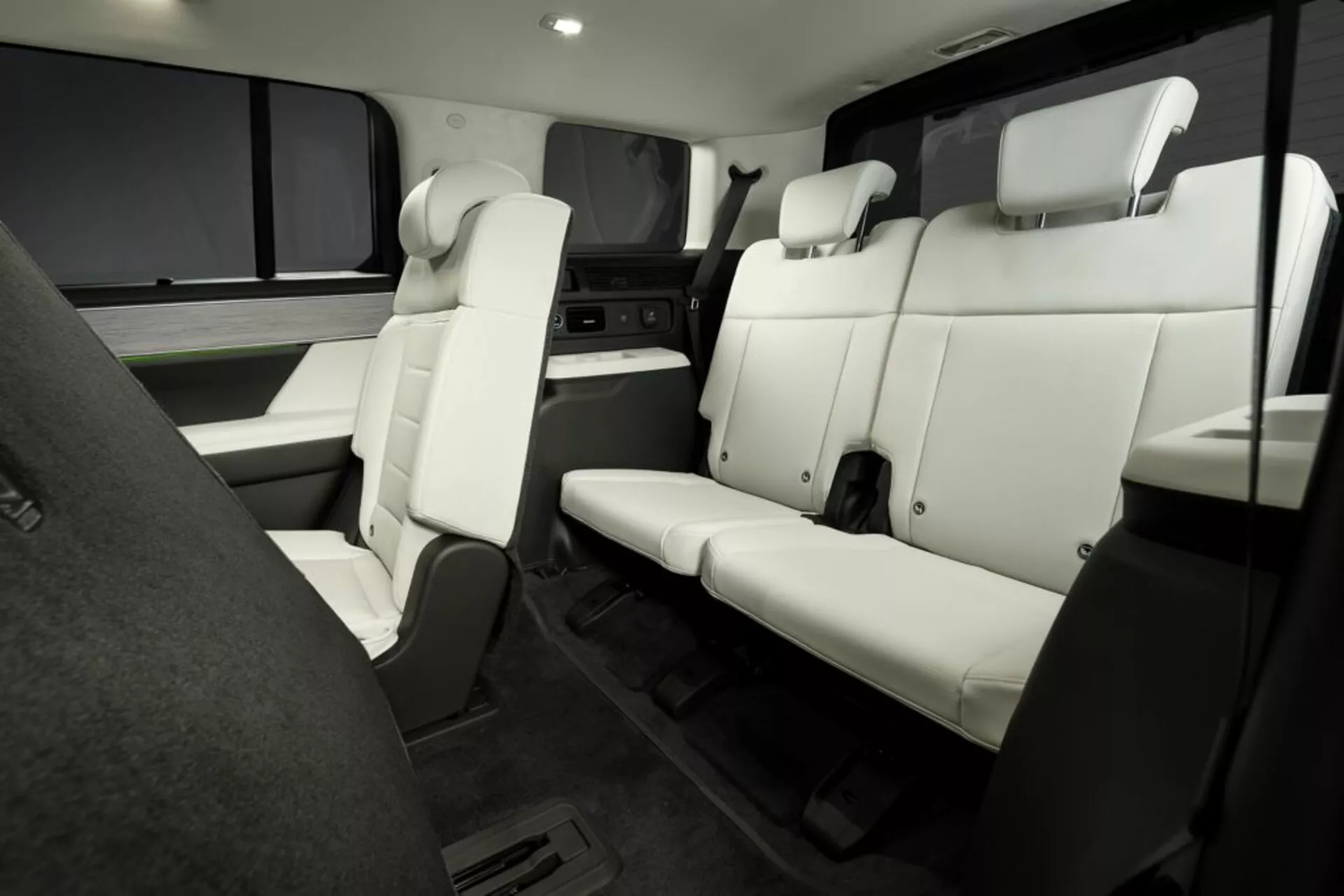 Tento snímek zobrazuje interiér moderního vozidla, konkrétně Hyundai Santa Fe 2024 (5. generace). Fotografie, pořízená z pohledu z předního sedadla, představuje boční pohled na uspořádání zadních sedadel. Sedadla, čalouněná bílou kůží nebo podobným materiálem, obsahují trojmístnou lavici s jasně definovanými opěrkami hlavy nejblíže k fotoaparátu. Za touto lavicí jsou vidět dvě částečně zakryté kapitánské židle s područkami. Uspořádání interiéru naznačuje prostornost a zaměření na luxus, s prvky jako rovná, nezakrytá podlaha a kryty oken. Snímek, dobře osvětlený a jasný, nevyzdvihuje žádnou špičkovou technologii ani elektronické ovládání.