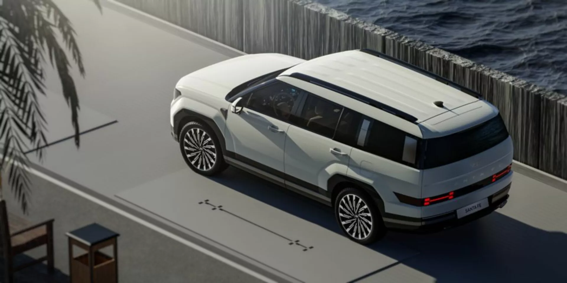 Pohled shora na SUV Hyundai Santa Fe páté generace 2024 zaparkované u vodní plochy, s dvoubarevným lakem v bílé a tmavém odstínu. Fotografie zobrazuje panoramatické střešní okno, velká vícepaprsková kola, opláštění karoserie a elegantní LED osvětlení. Scéna je klidná, s přirozeným osvětlením naznačujícím buď ráno nebo odpoledne, a naznačuje luxusní nebo prémiové prostředí s potenciálními pokročilými technologickými funkcemi, jako jsou senzory blízkosti a senzory okolního světla.